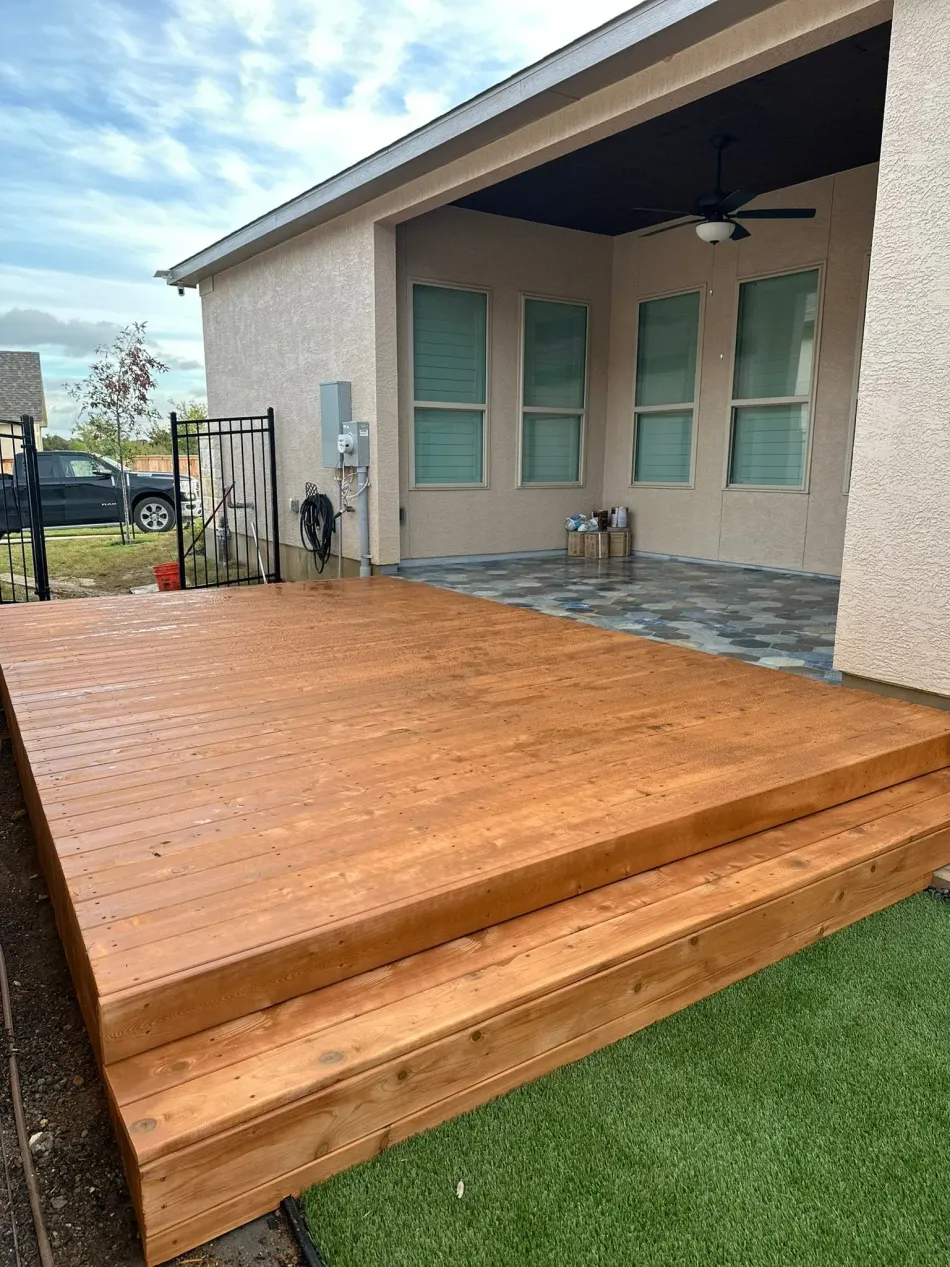 Wooden deck with steps next to a house with beige stucco walls, next to artificial green grass.
