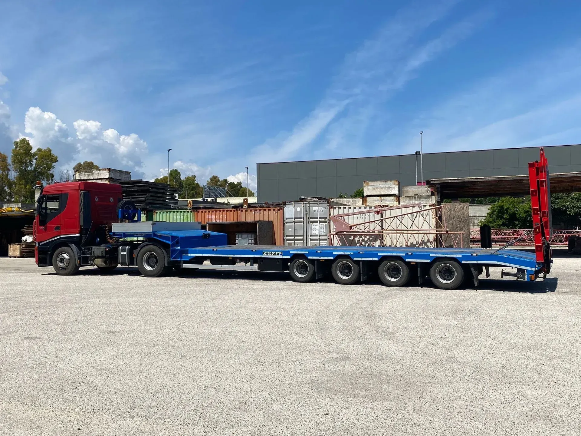 Un camion rosso che traina un lungo rimorchio a pianale blu in un piazzale ghiaioso sotto un cielo azzurro.