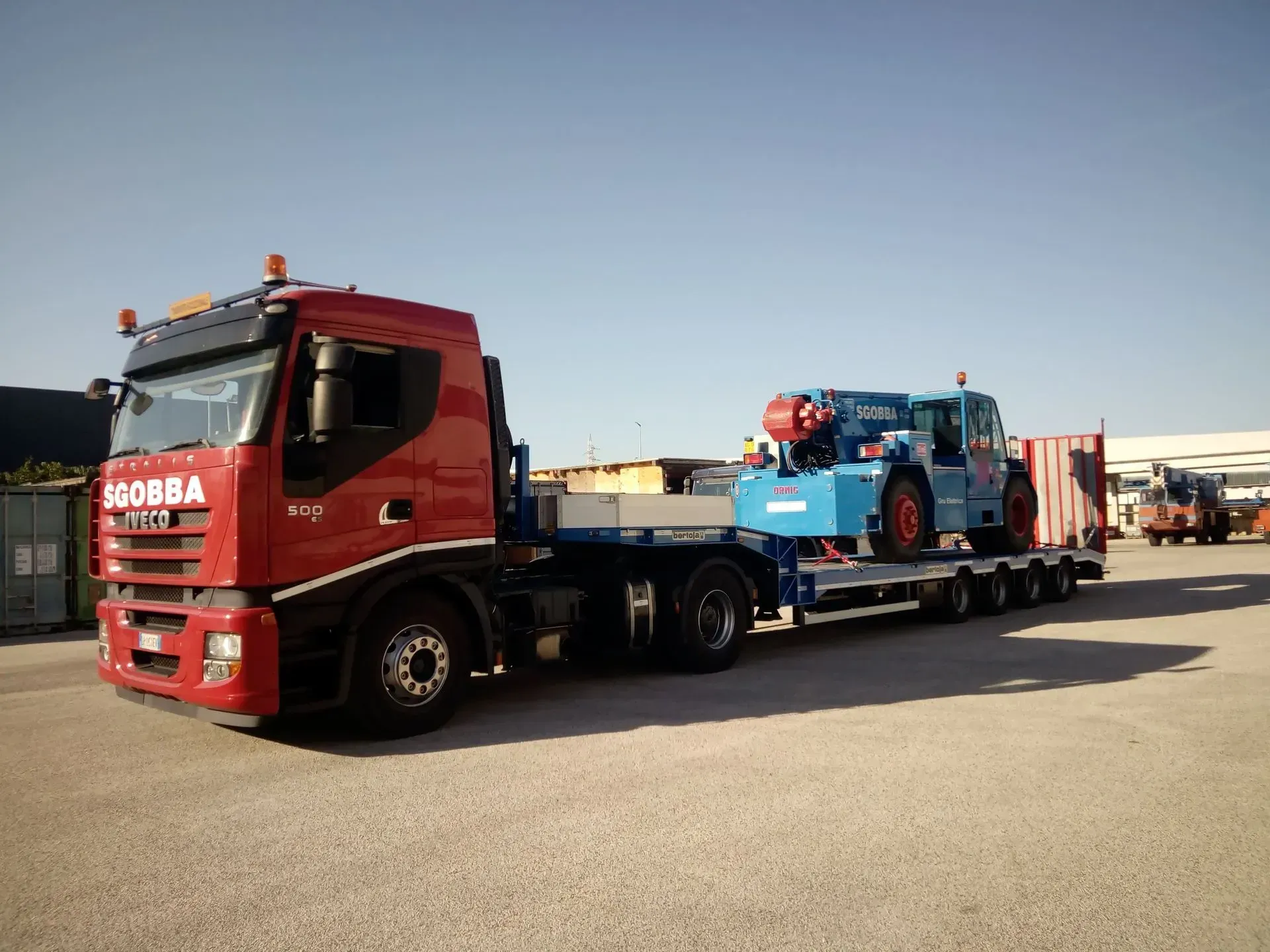 Un camion rosso che trasporta un'auto da corsa blu su un rimorchio a pianale in un parcheggio.