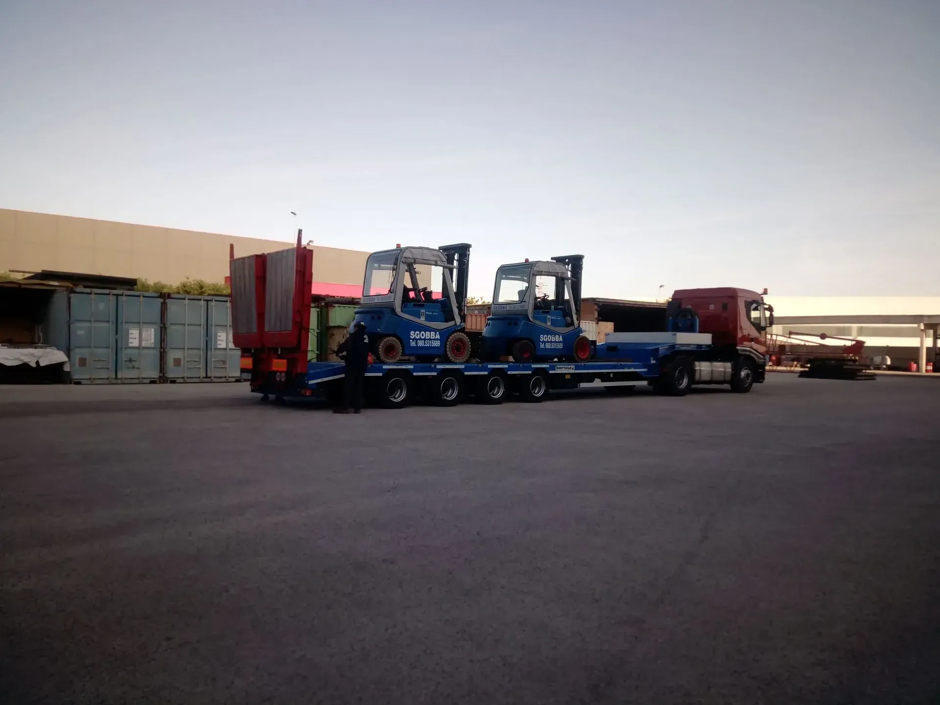 Un semirimorchio che trasporta carrelli elevatori blu su un rimorchio a pianale in un parcheggio al tramonto.