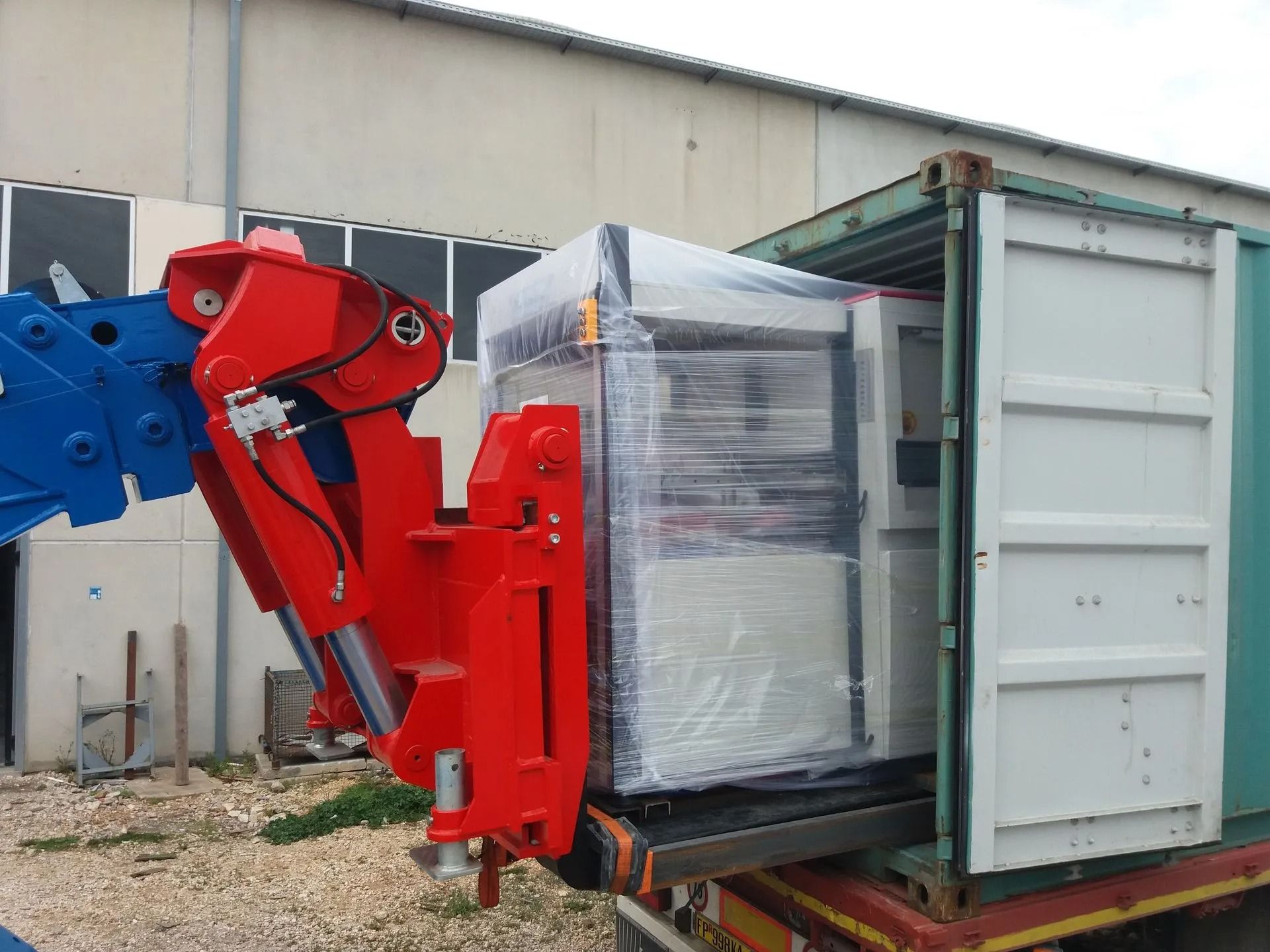 Macchinari idraulici rossi sollevano un pallet avvolto in pellicola termoretraibile e lo inseriscono in un container aperto.