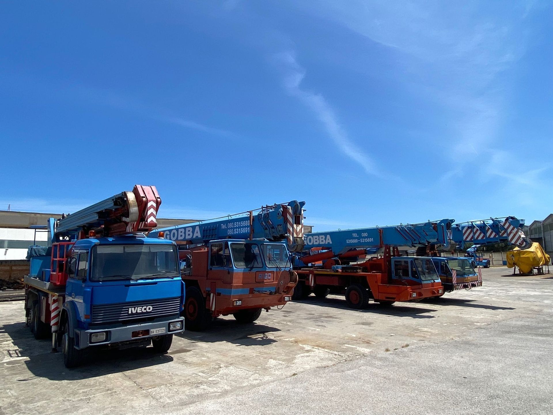 Carri attrezzi blu e arancioni parcheggiati in un cortile sotto un cielo azzurro brillante