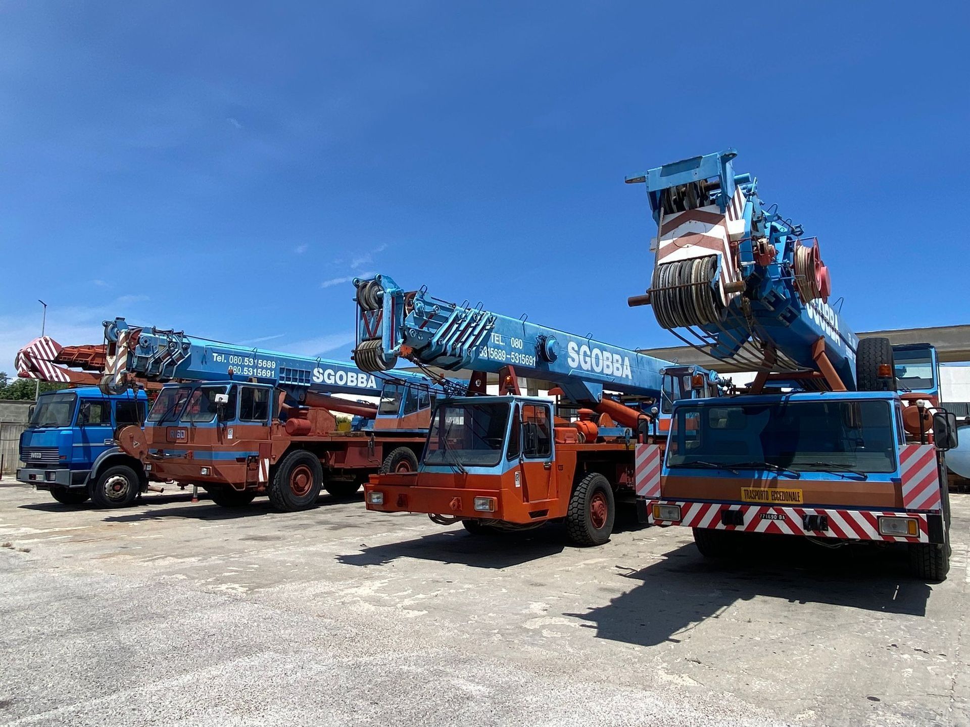 Una fila di camion gru blu e arancioni parcheggiati in un piazzale sotto un cielo sereno