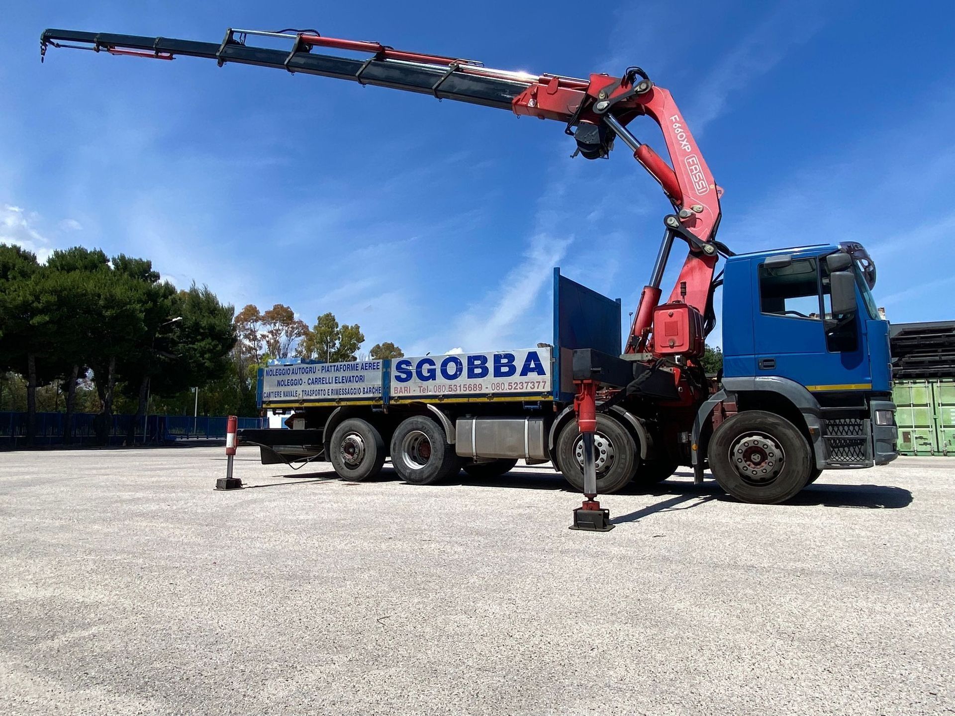 In un soleggiato piazzale industriale, una gru rossa montata su un camion solleva un lungo braccio sopra un rimorchio bianco.