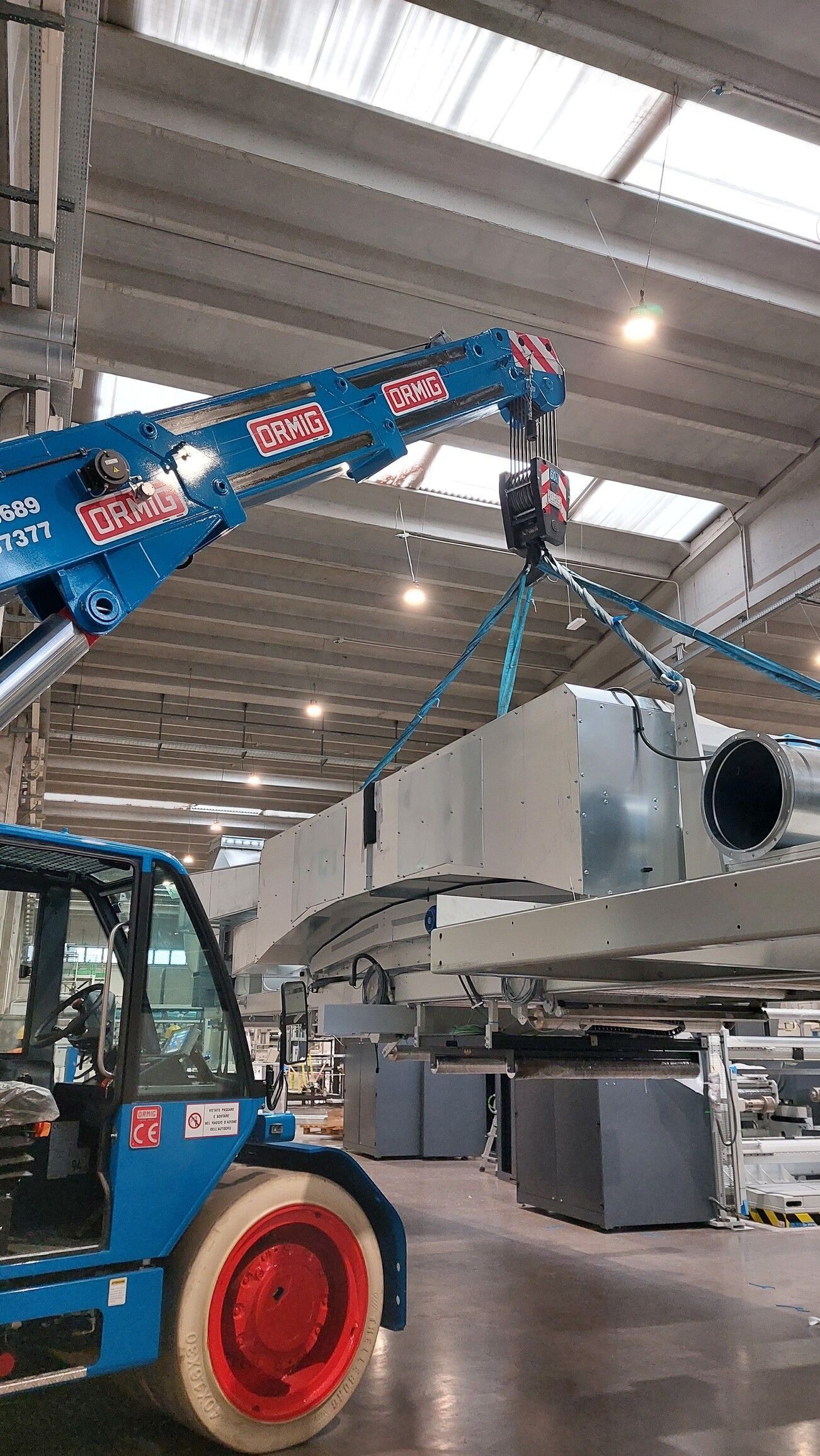 Un carrello elevatore blu solleva una grande unità HVAC metallica all'interno di un magazzino con soffitti alti.