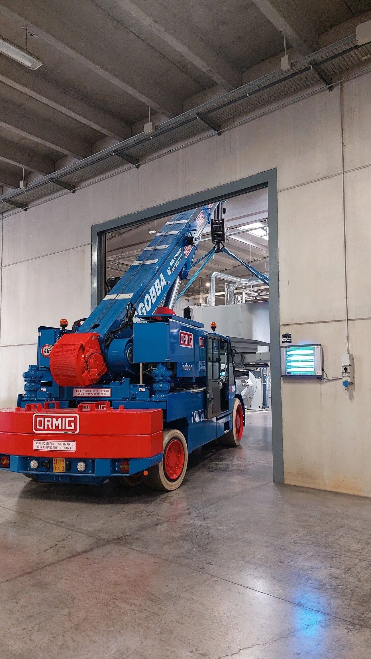 Piattaforma elevatrice mobile blu e rossa parcheggiata all'interno di un garage industriale, con il braccio sollevato verso una porta.