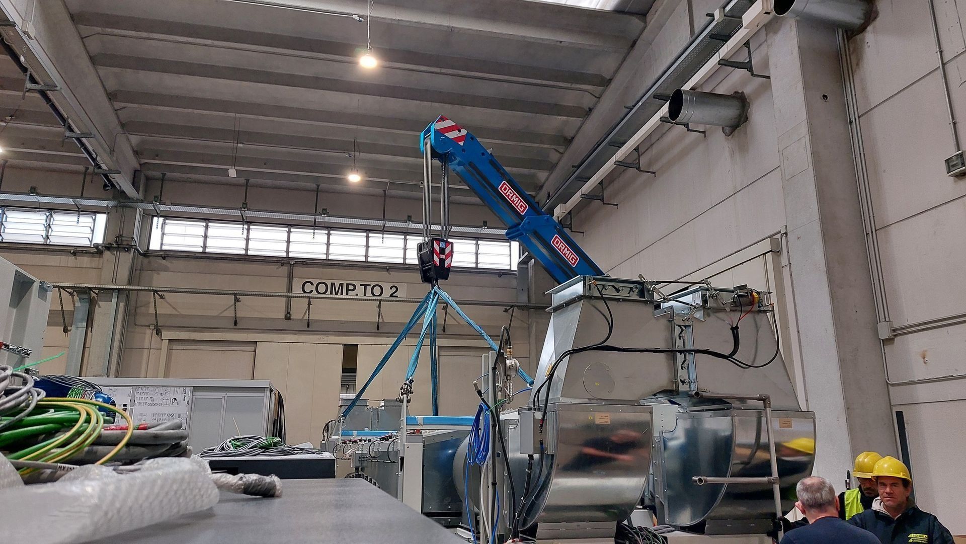 Magazzino industriale con una gru blu che solleva attrezzature sotto un soffitto alto, operai nelle vicinanze.