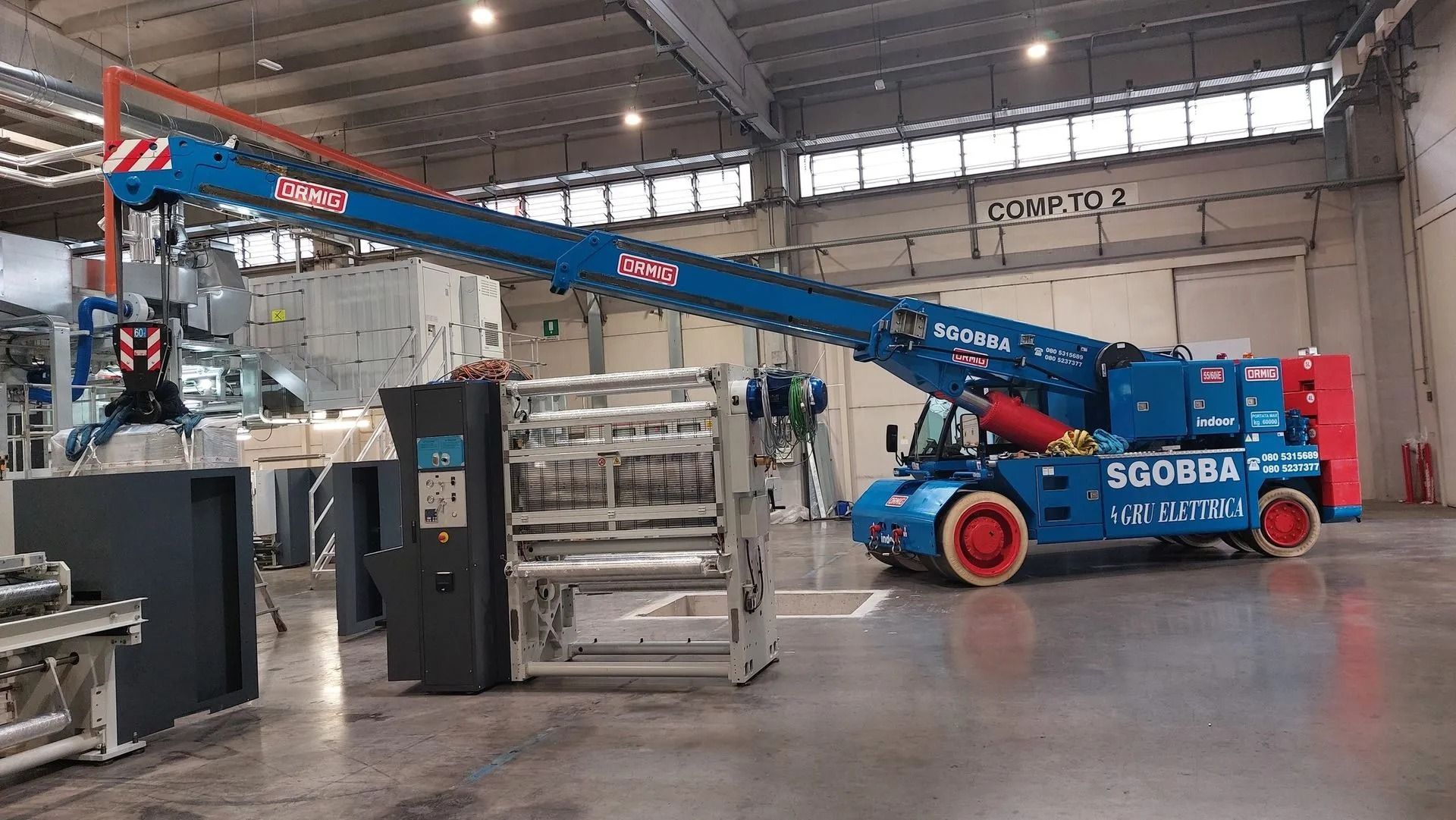 Piattaforma aerea telescopica blu e rossa in un magazzino, accanto a pallet e attrezzature impilati.