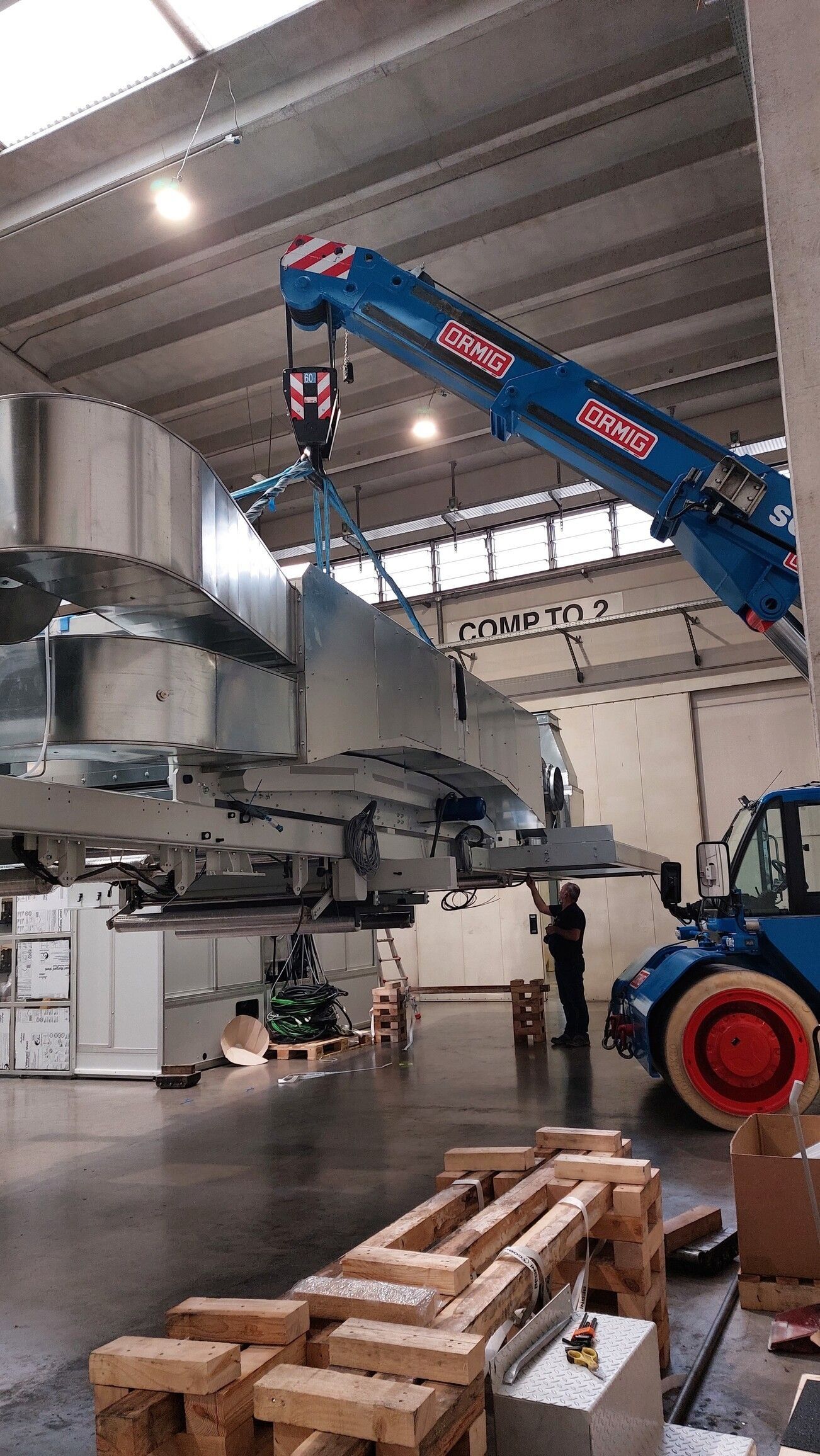 Una gru blu solleva un grande componente grigio di un aereo in un magazzino industriale vicino a pallet di legno.