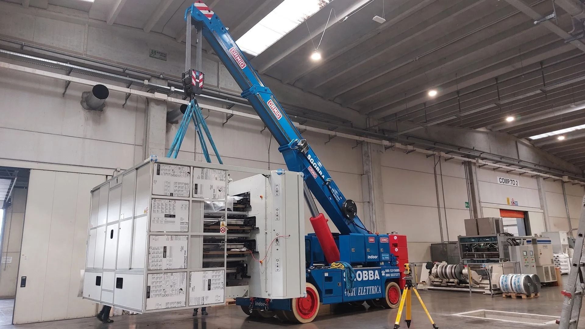 Una gru blu solleva un grande macchinario bianco all'interno di un magazzino industriale.