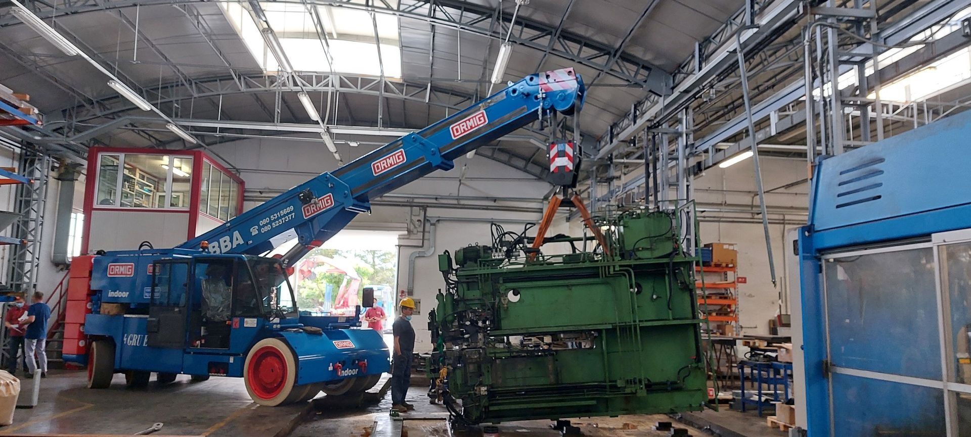 Una gru blu solleva un macchinario industriale verde all'interno di un'officina di fabbrica.