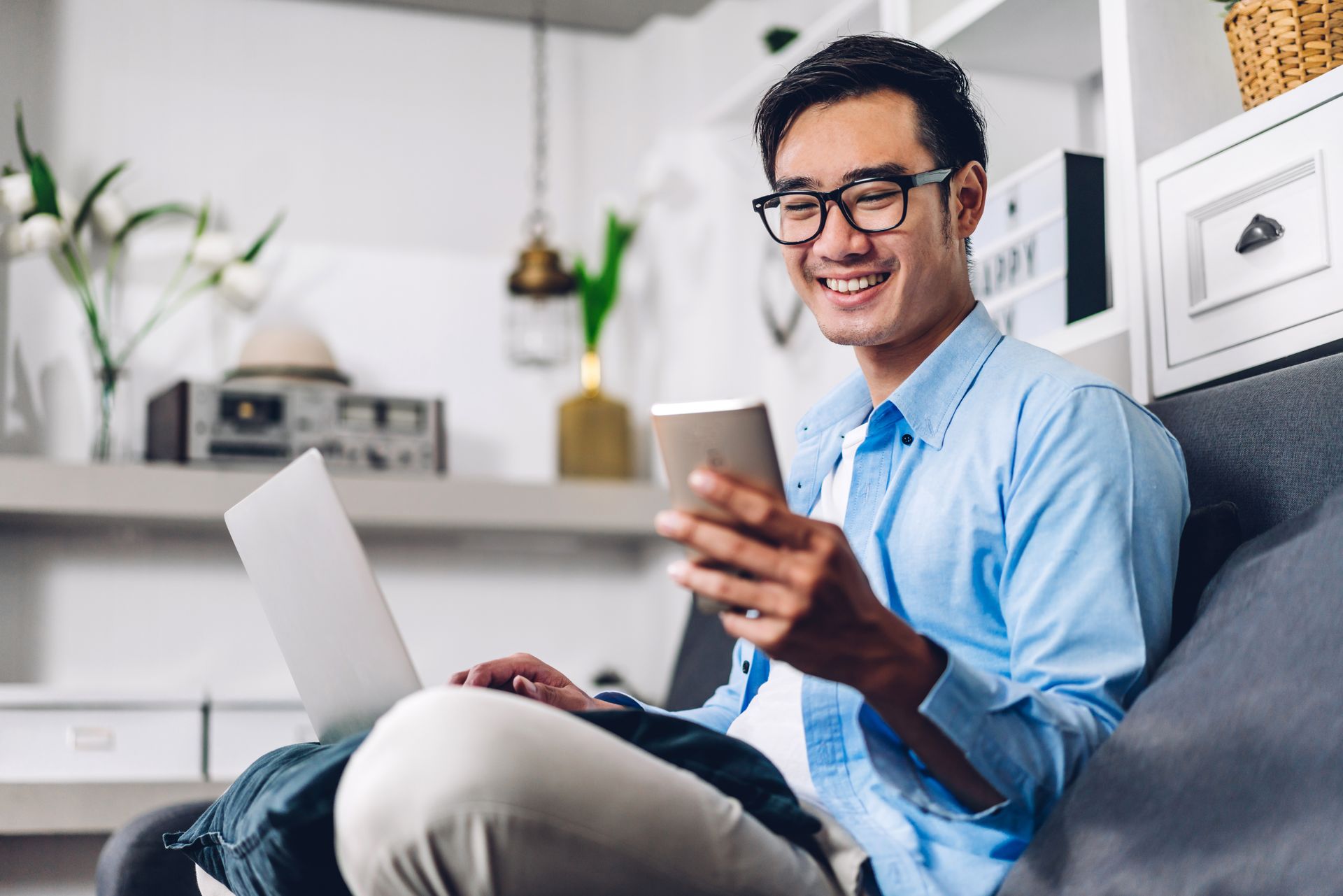 A man is sitting on a couch using a laptop and a cell phone.