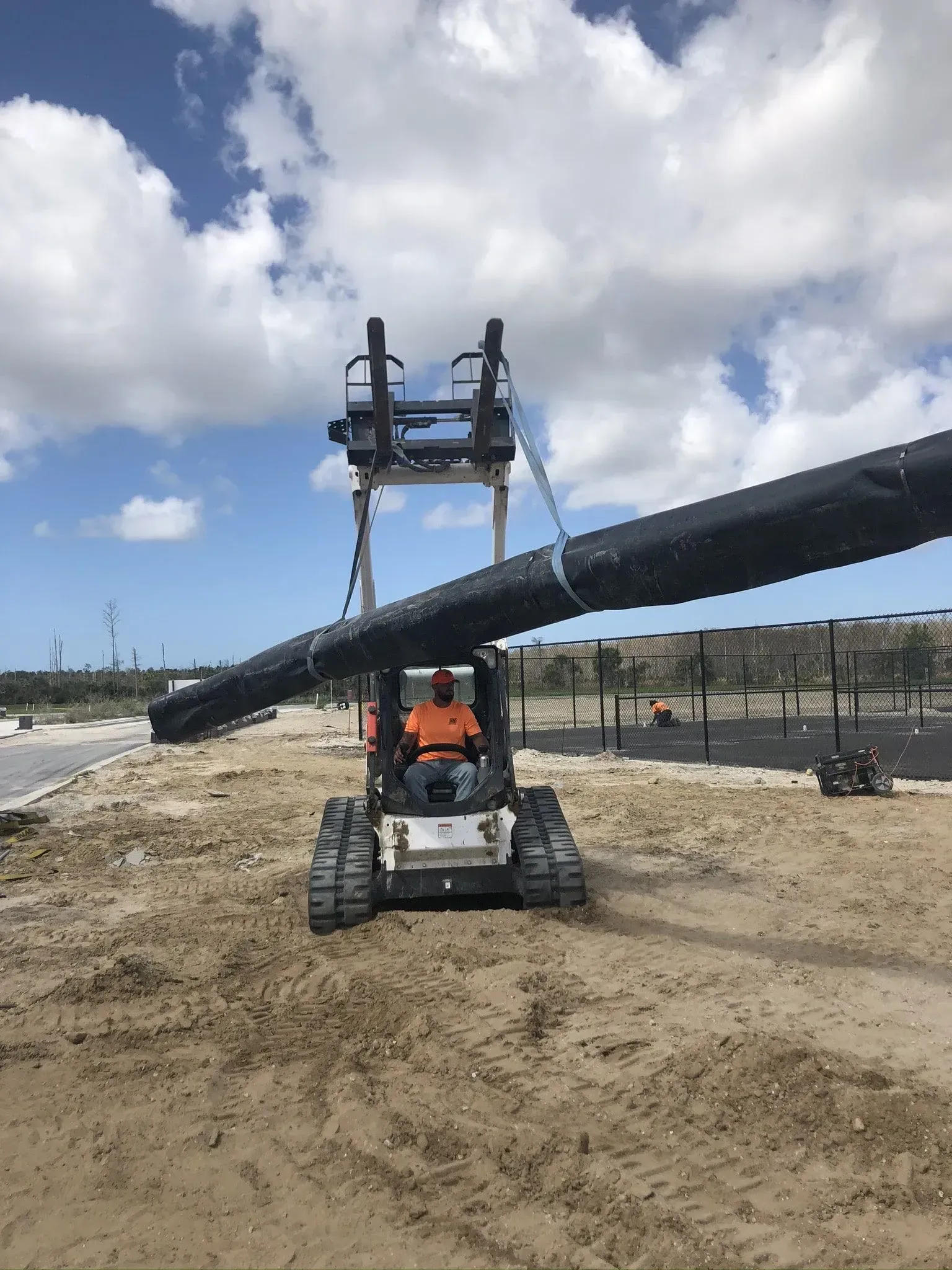 Worker on a lift installing a large black pipe at a construction site under a cloudy sky