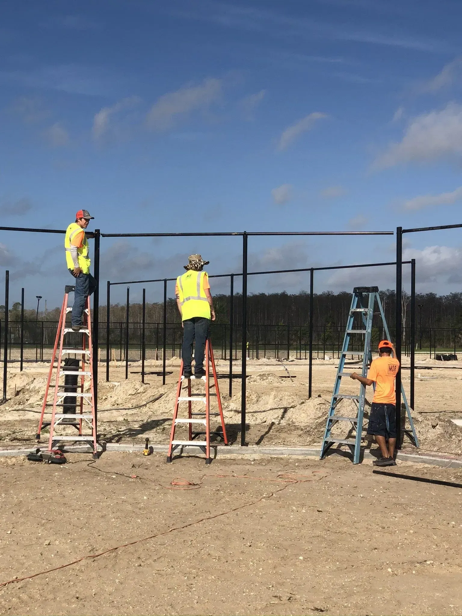 Workers on ladders installing a black metal fence in a dirt field under a blue sky