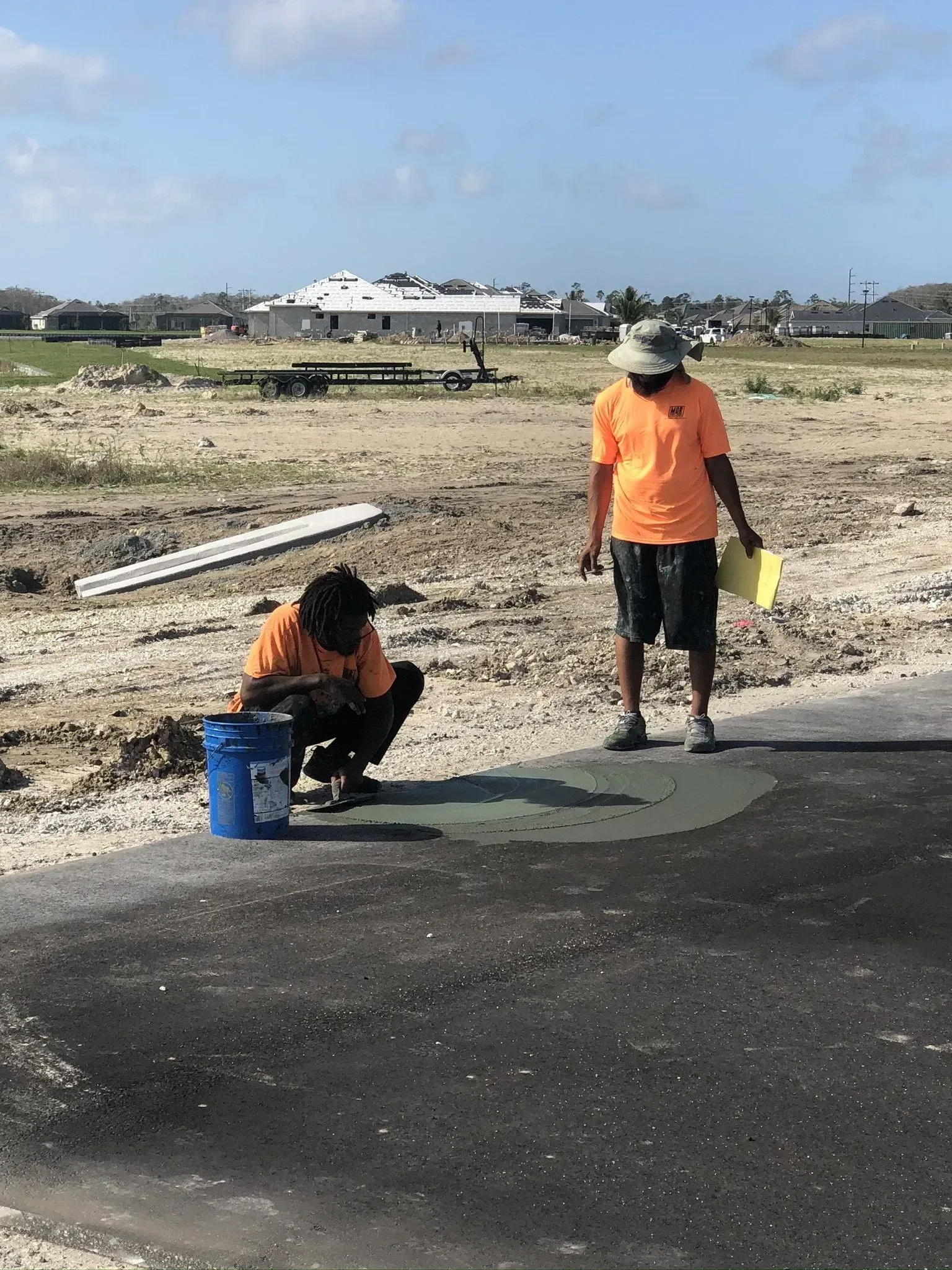 Two workers in orange shirts repairing a paved road in a dusty open area.