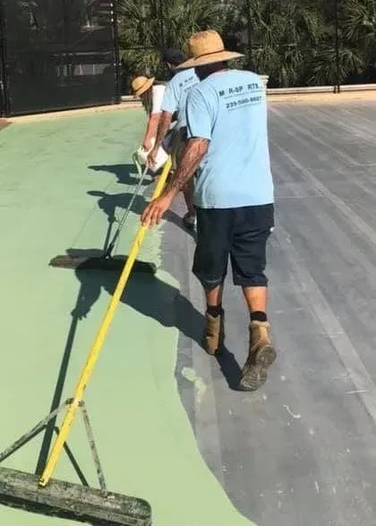 Two workers painting a large outdoor surface with a yellow pole and roller, green paint beside gray pavement