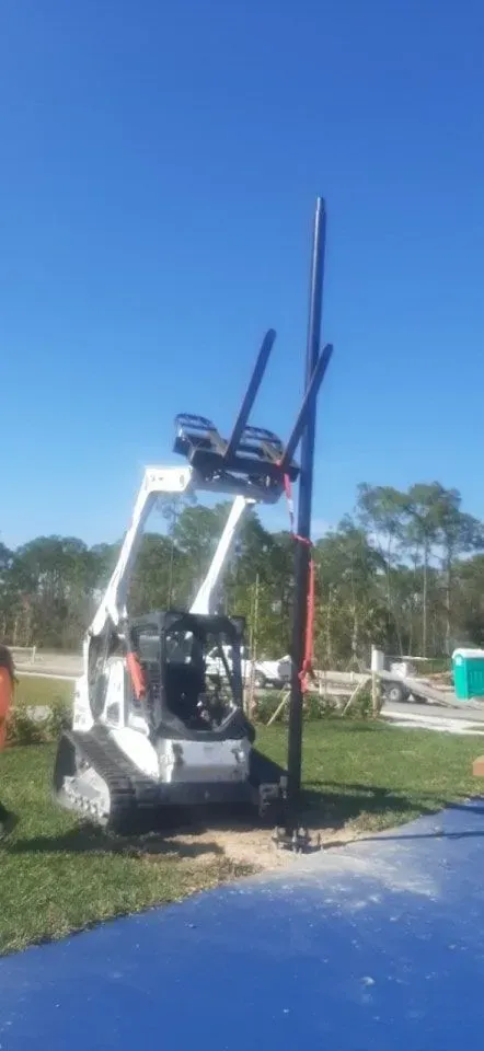 White forklift lifting a tall black pole beside a blue surface on a sunny roadside.