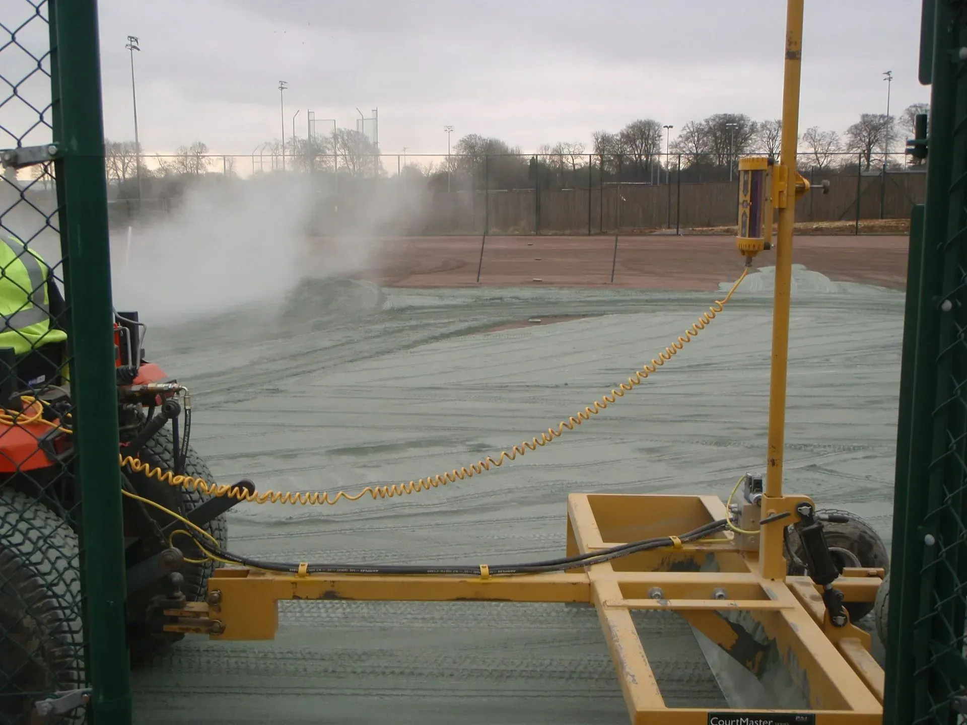 Yellow field machine spraying mist on a tarp-covered surface near a chain-link fence and sports facility.