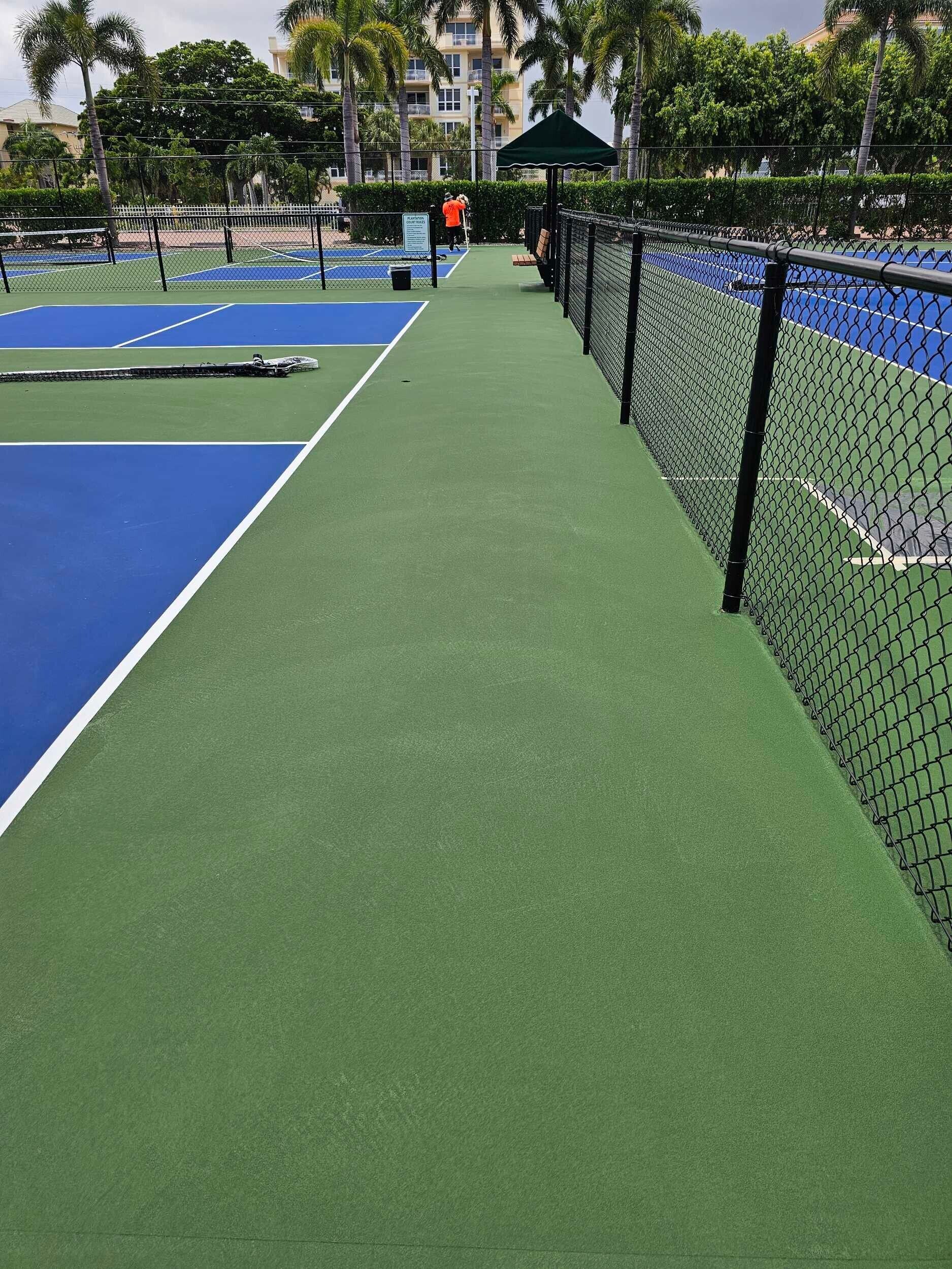 Outdoor tennis courts beside a green walkway and black fence, with palm trees and buildings in the background