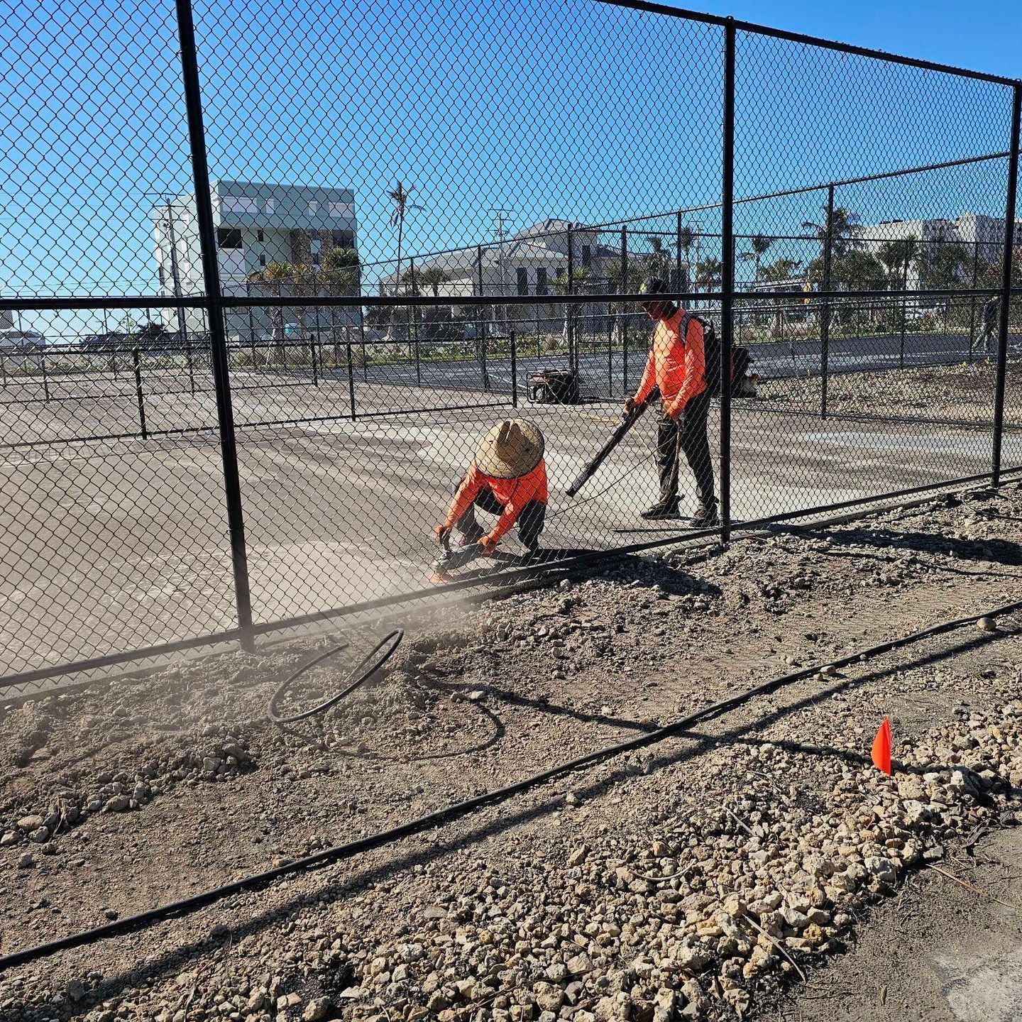 Two workers in orange safety gear repairing a fence beside a gravel area under a clear blue sky