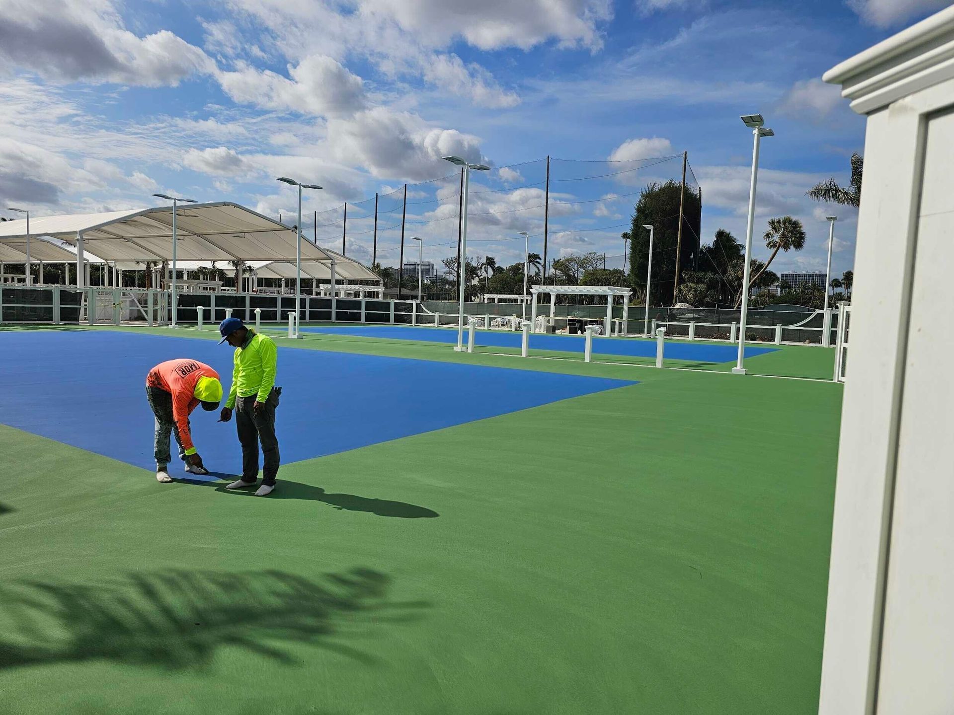Two workers marking lines on a blue and green pickleball court under a cloudy sky