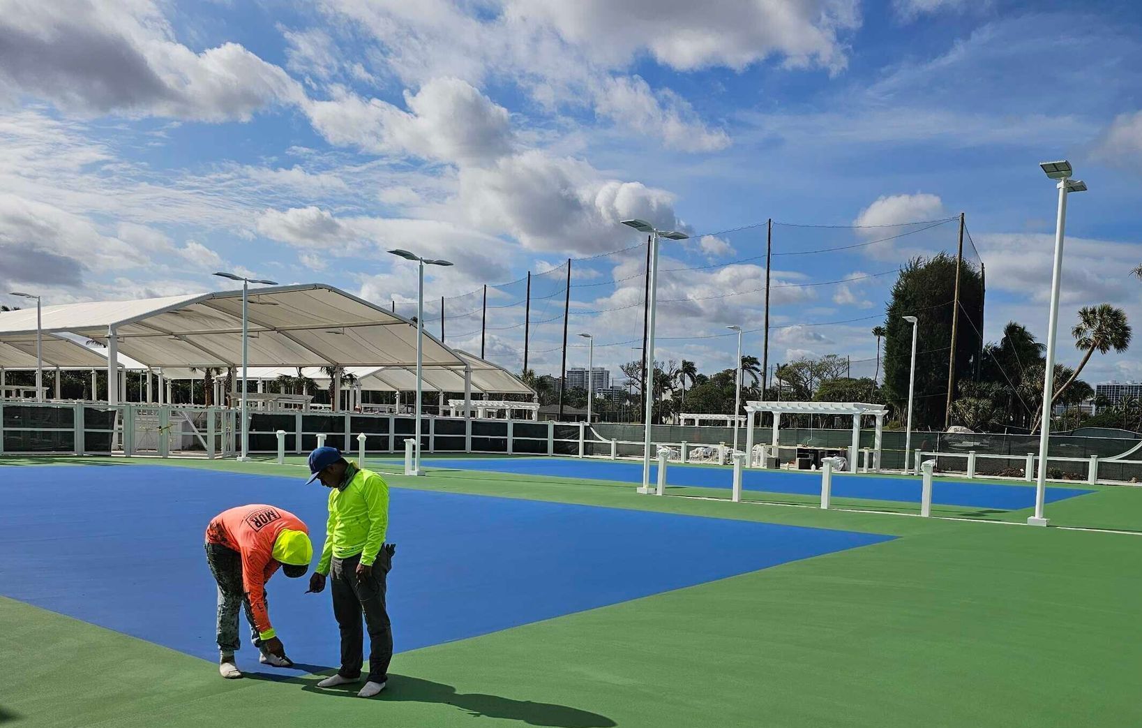 Two workers marking a blue outdoor court on a green field under a cloudy sky