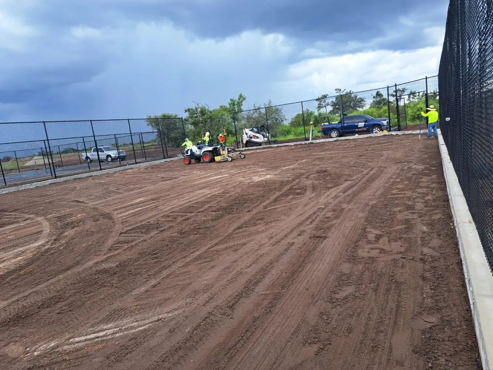 Dirt sports field with a few workers and equipment near a fence under a cloudy sky