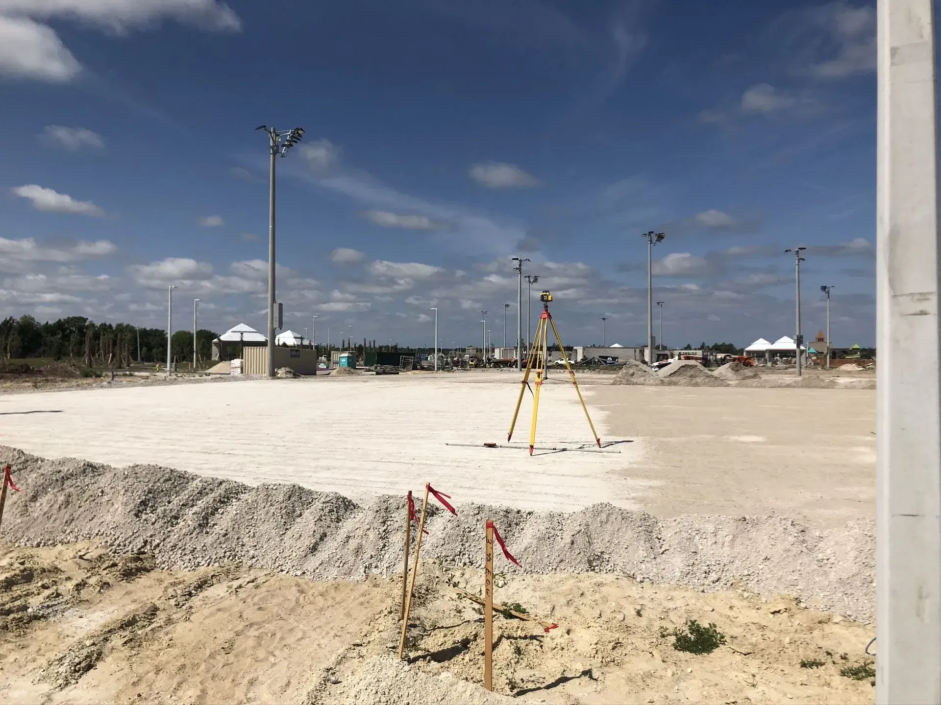 Construction site with survey equipment, sandy ground, and tall light poles under a blue sky