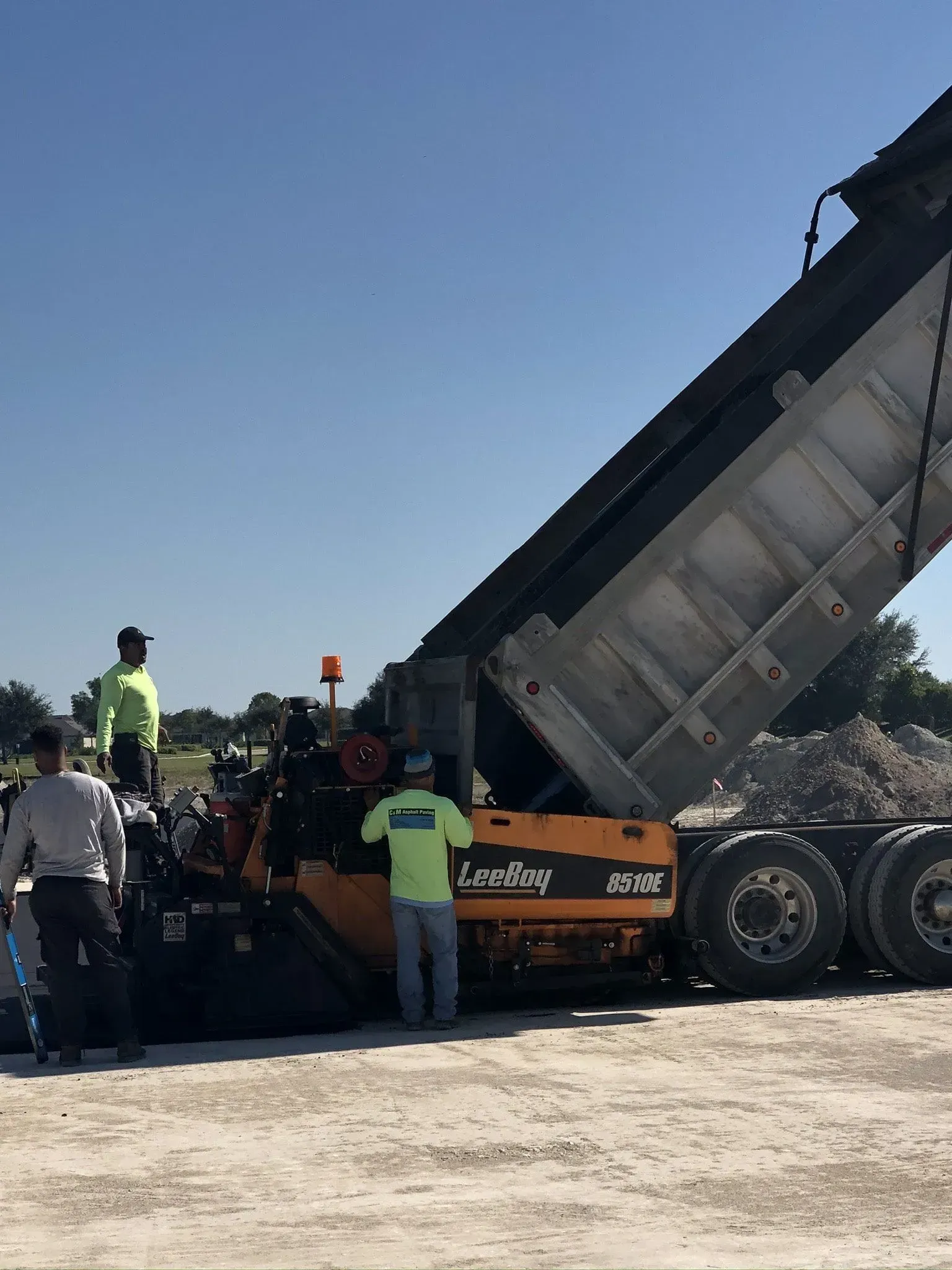 Workers operate a dump truck unloading dirt at a construction site under a clear sky.