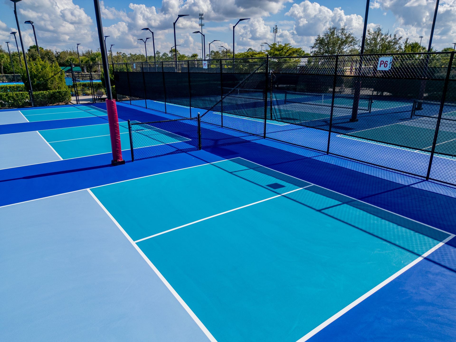 Blue outdoor pickleball courts with black fencing and netted divider under a cloudy sky