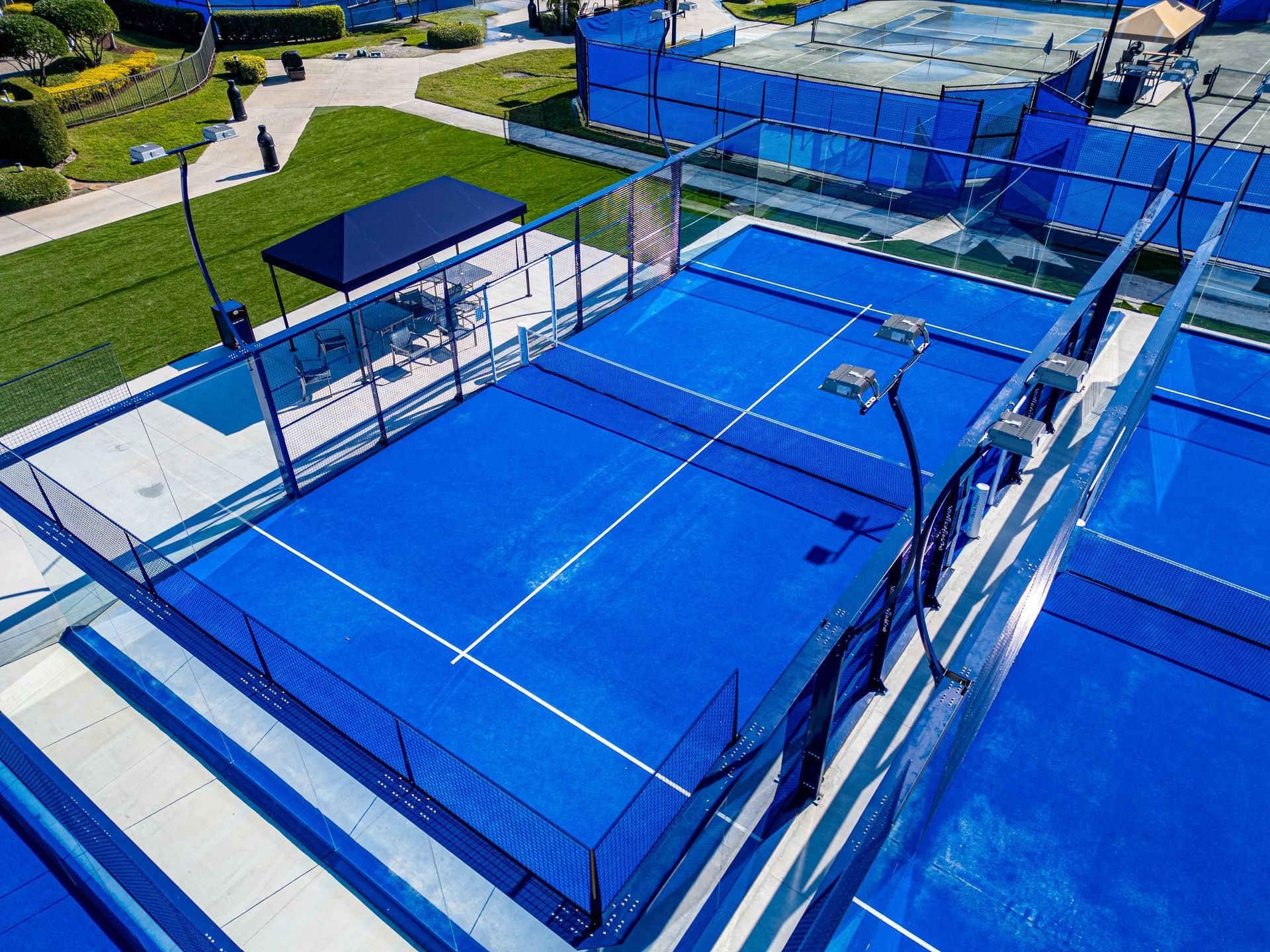 Aerial view of a blue outdoor tennis court with nets, fences, and surrounding walkways.