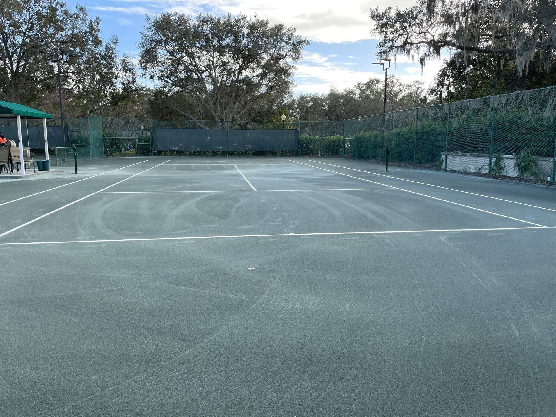Empty outdoor tennis court with white lines and a fence, surrounded by trees under a cloudy sky