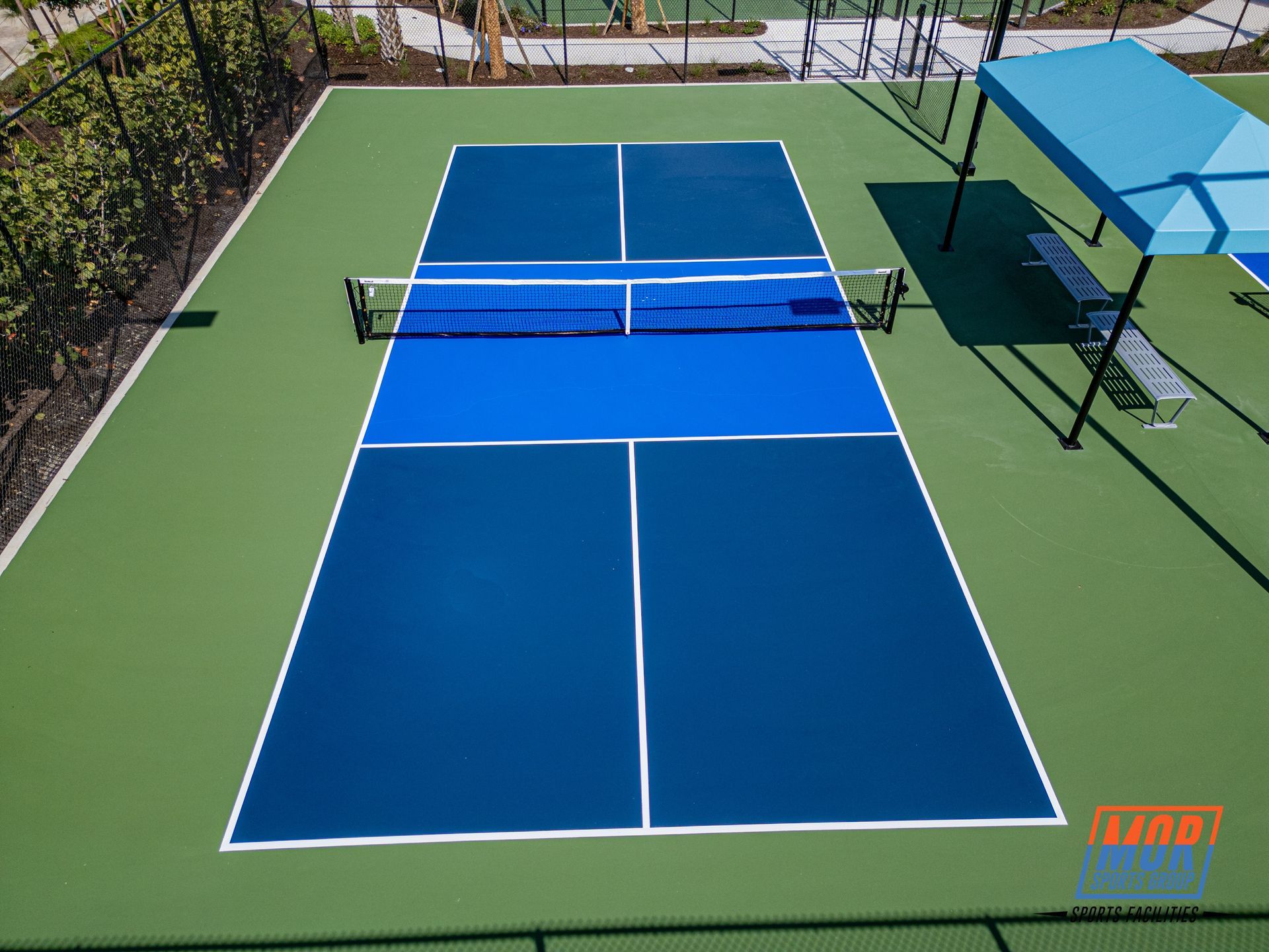 Blue tennis court with a net, surrounded by green fencing and palm trees under a clear sky