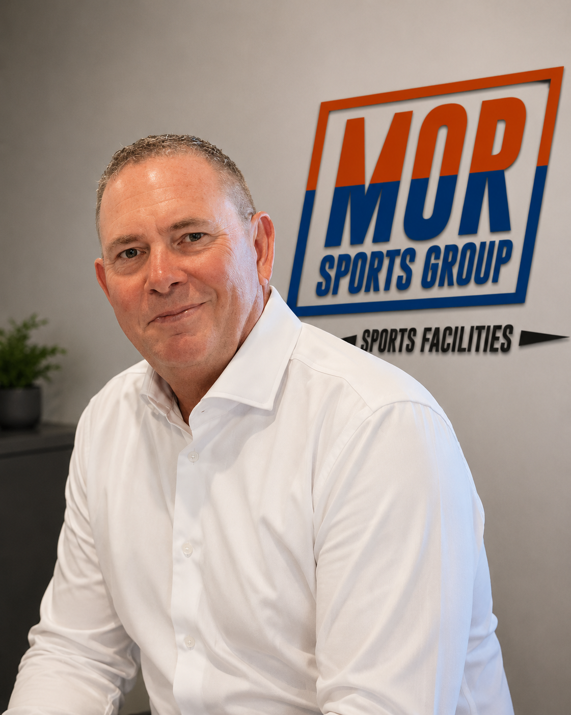Gary Morton - Man in a white shirt standing beside a MOR Sports Group logo in an office setting