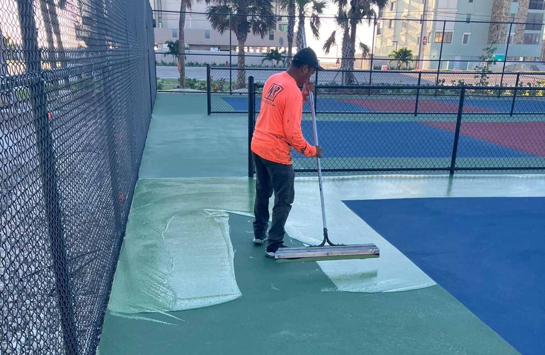Person in orange jacket cleaning a tennis court with a push squeegee near the net
