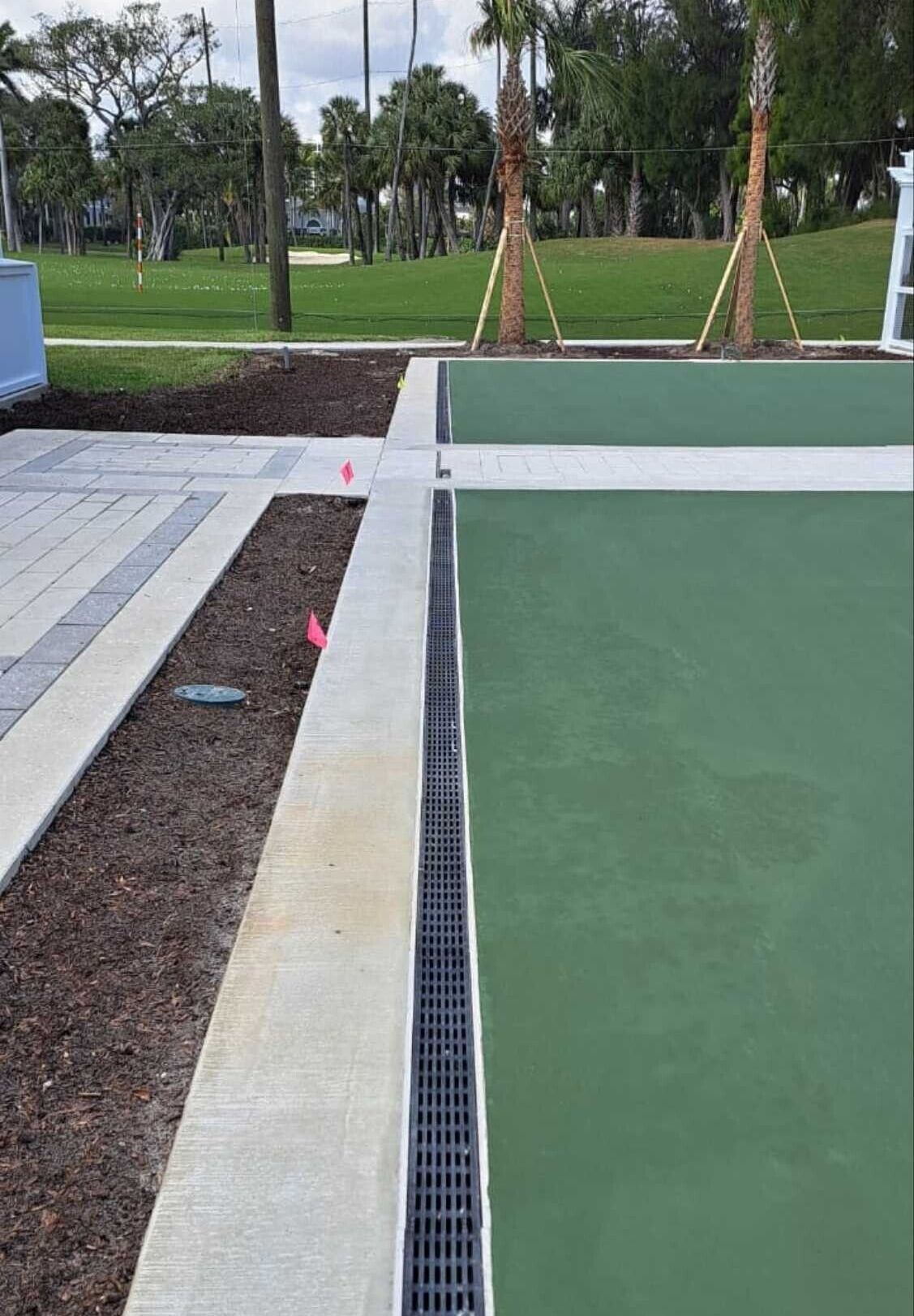 Outdoor court with a drainage channel separating green playing surface from a paved walkway and landscaped area.