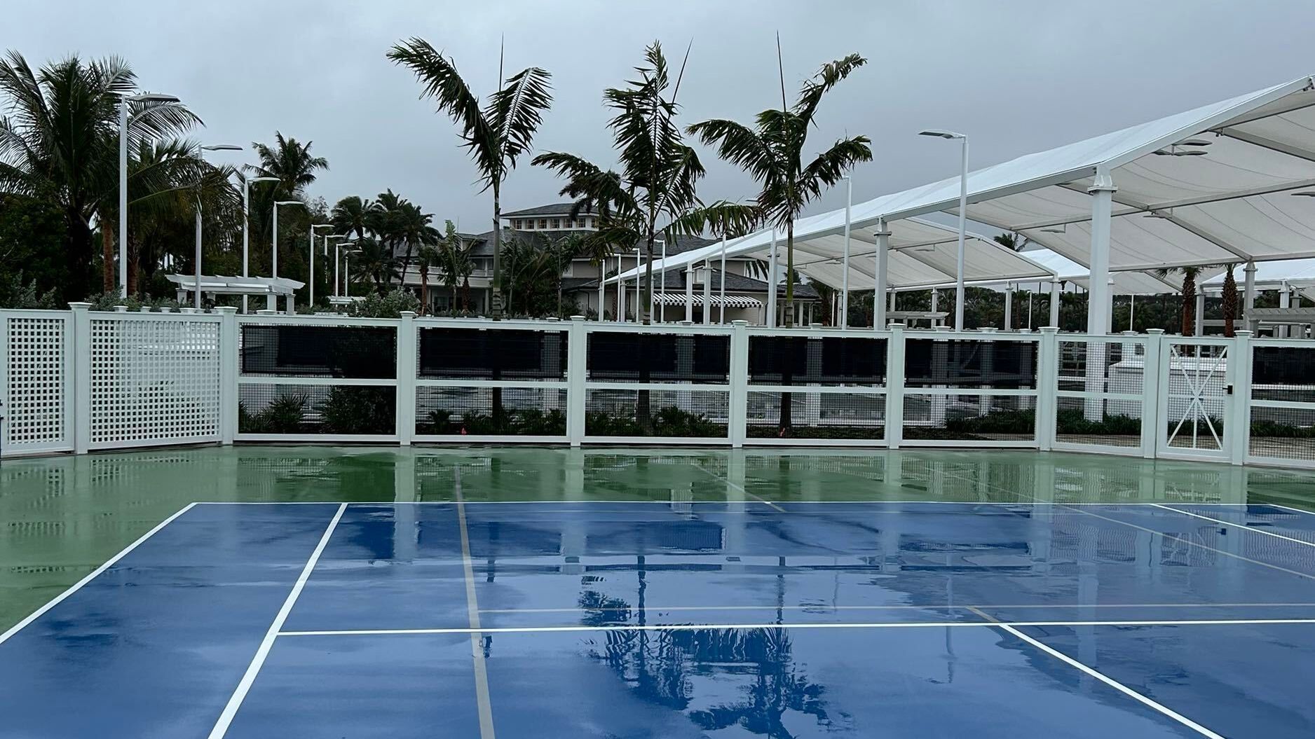Blue outdoor tennis court under cloudy skies, with palm trees and white pergola structures in the background