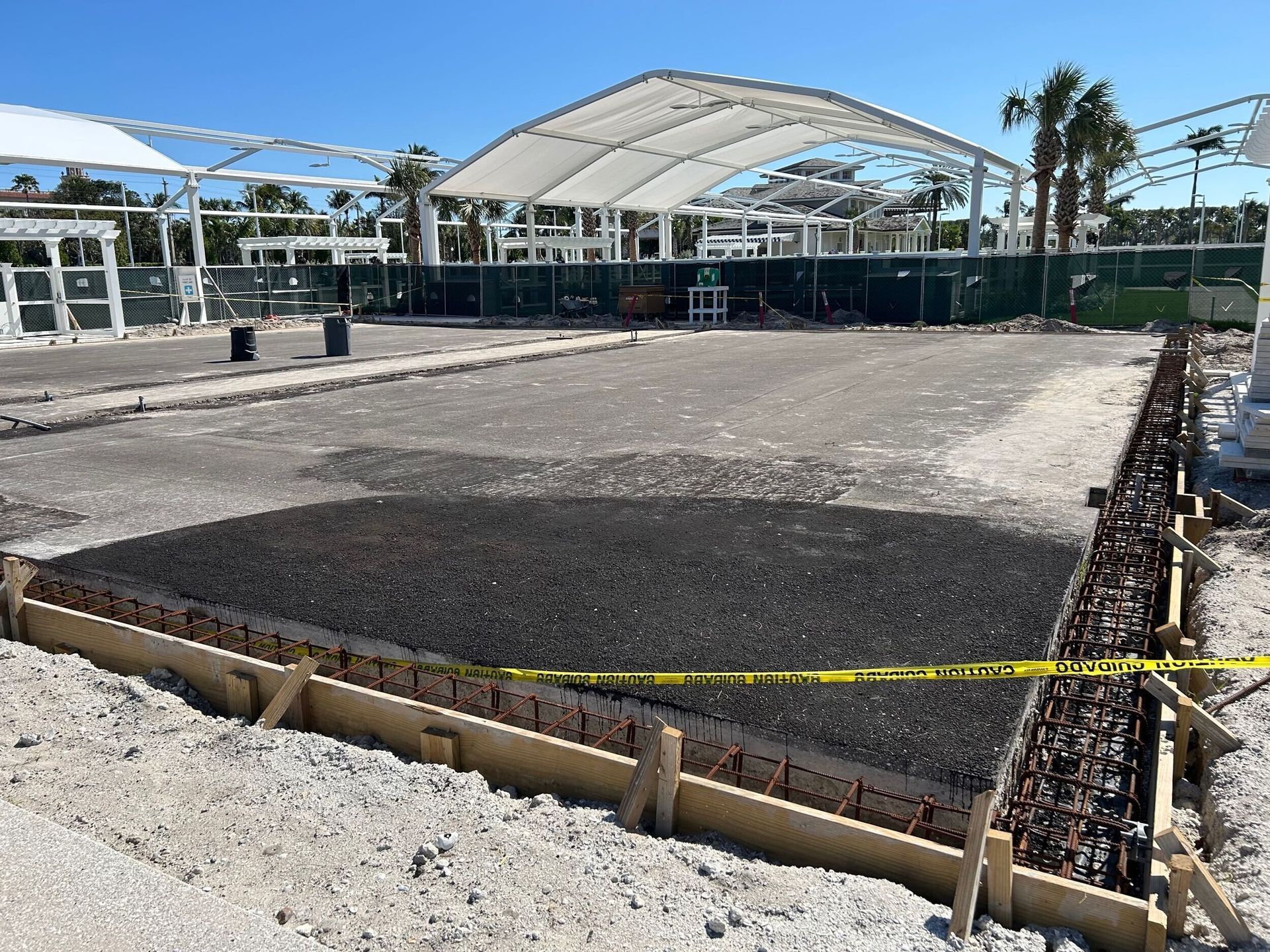 Empty construction site with gravel foundation, yellow caution tape, and white canopy structures in the background