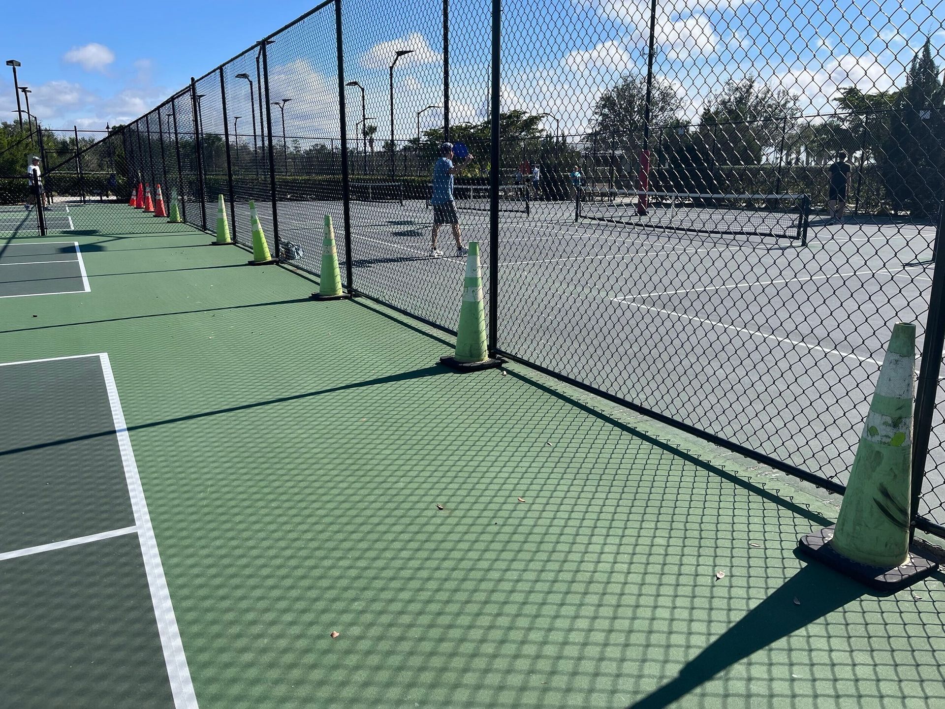 Empty outdoor pickleball court beside a chain-link fence, with green cones and a clear blue sky.