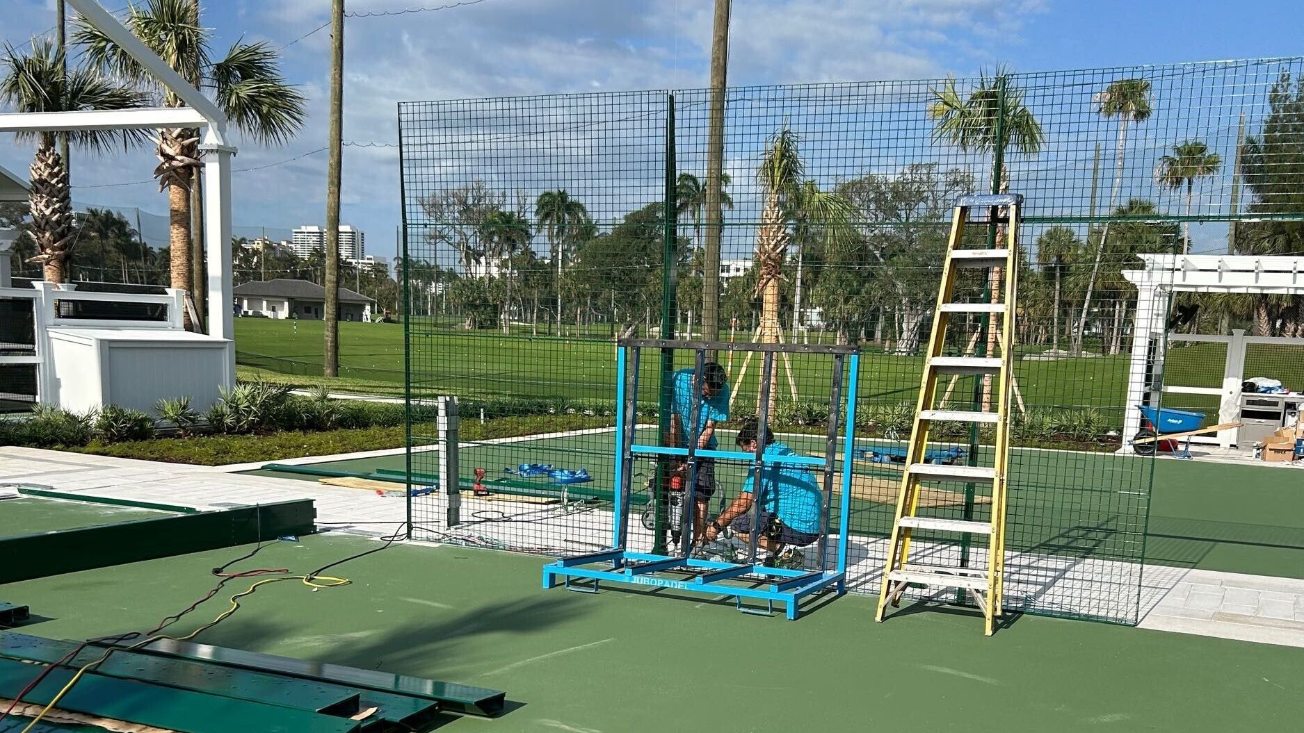 Blue exercise equipment and a yellow ladder on a green outdoor court, with a wire fence and trees behind.
