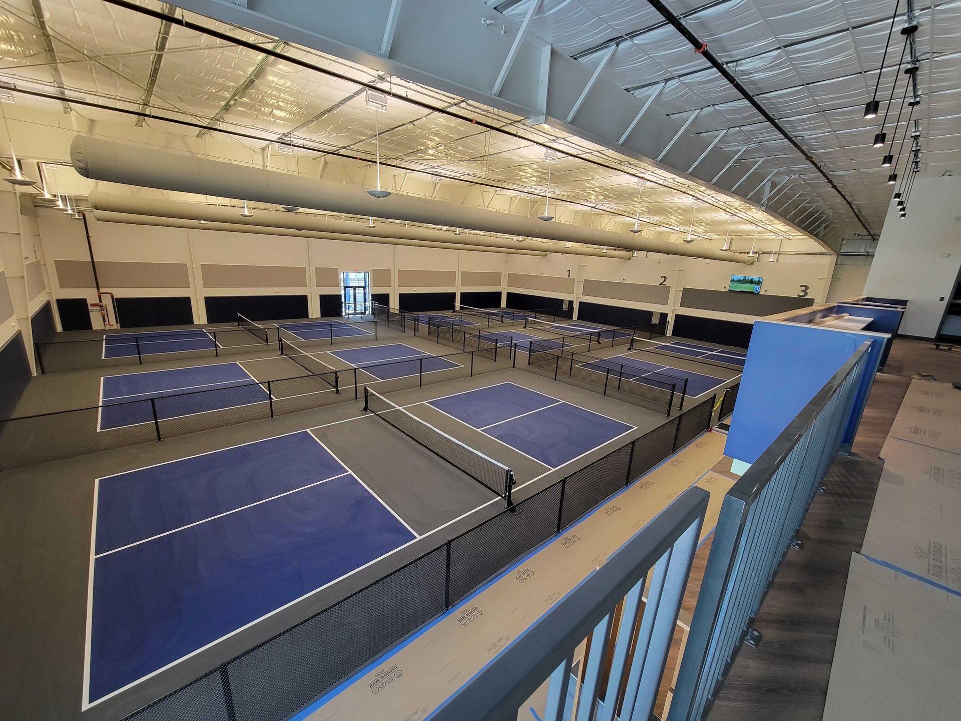 Indoor tennis courts with blue surfaces under a high arched ceiling, viewed from an upper balcony
