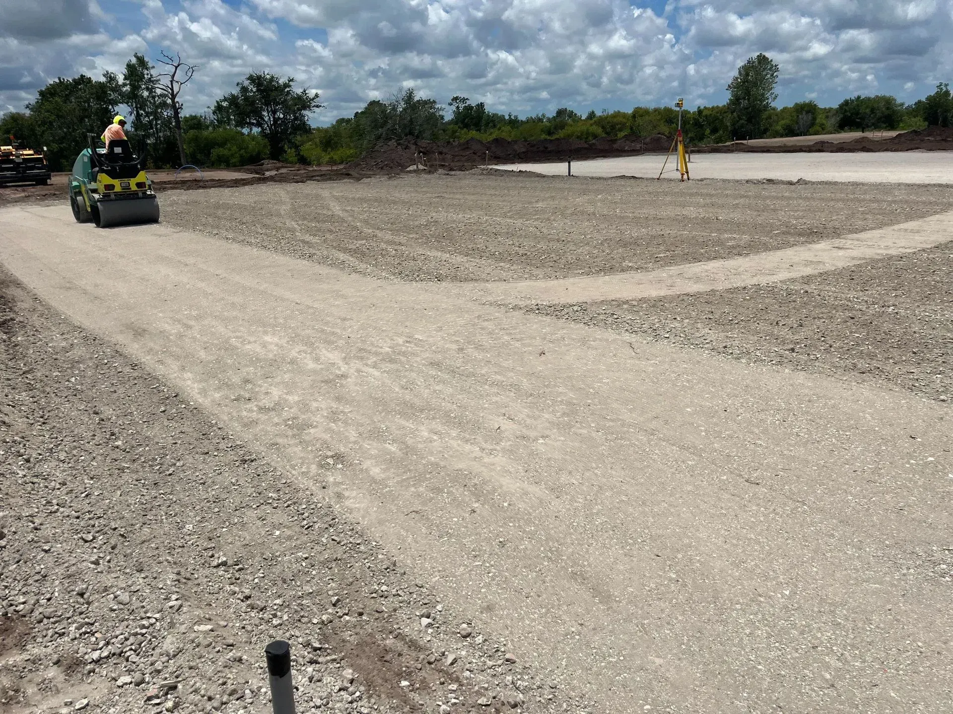 Two workers operate a small roller on a wide gravel road under a cloudy sky.