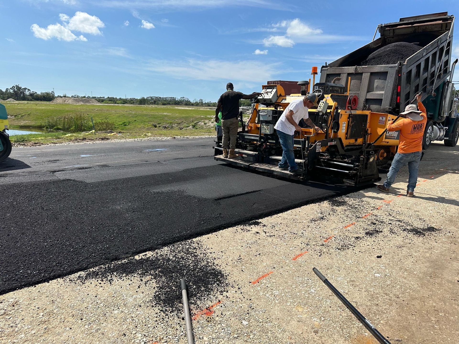 Road crew paving a rural road with black asphalt beside a dump truck on a sunny day