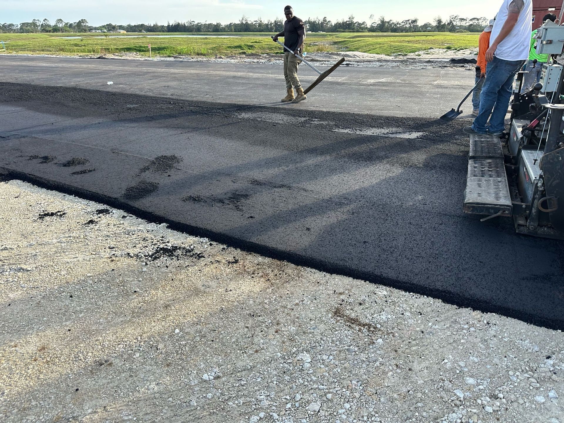 Road workers paving asphalt on a rural road, with a roller and fresh black pavement beside gravel.