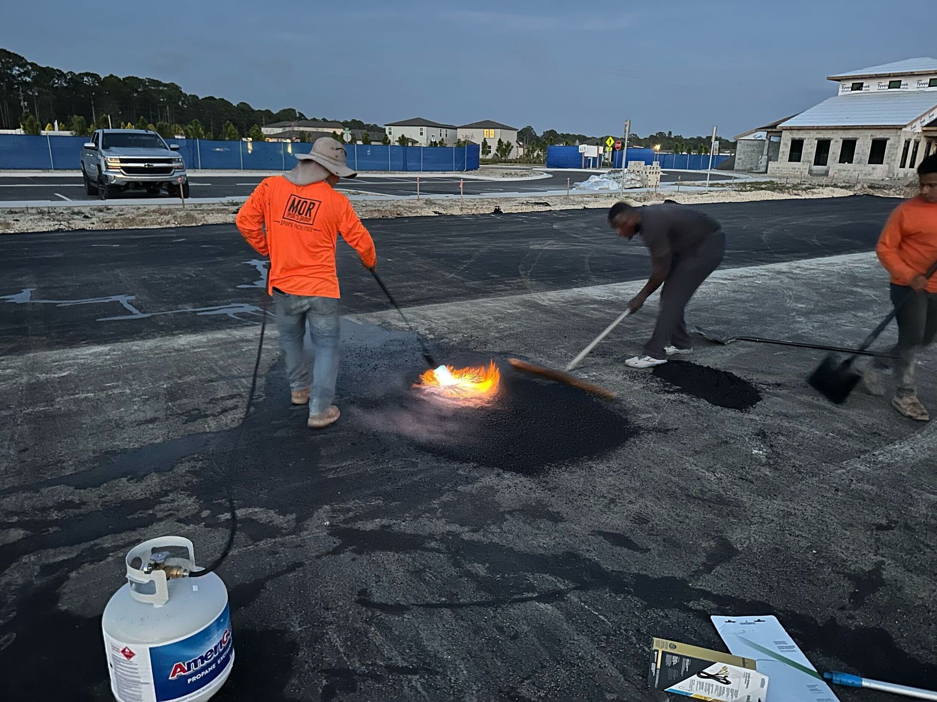 Workers patching hot asphalt on a paved surface near parked trucks and buildings.