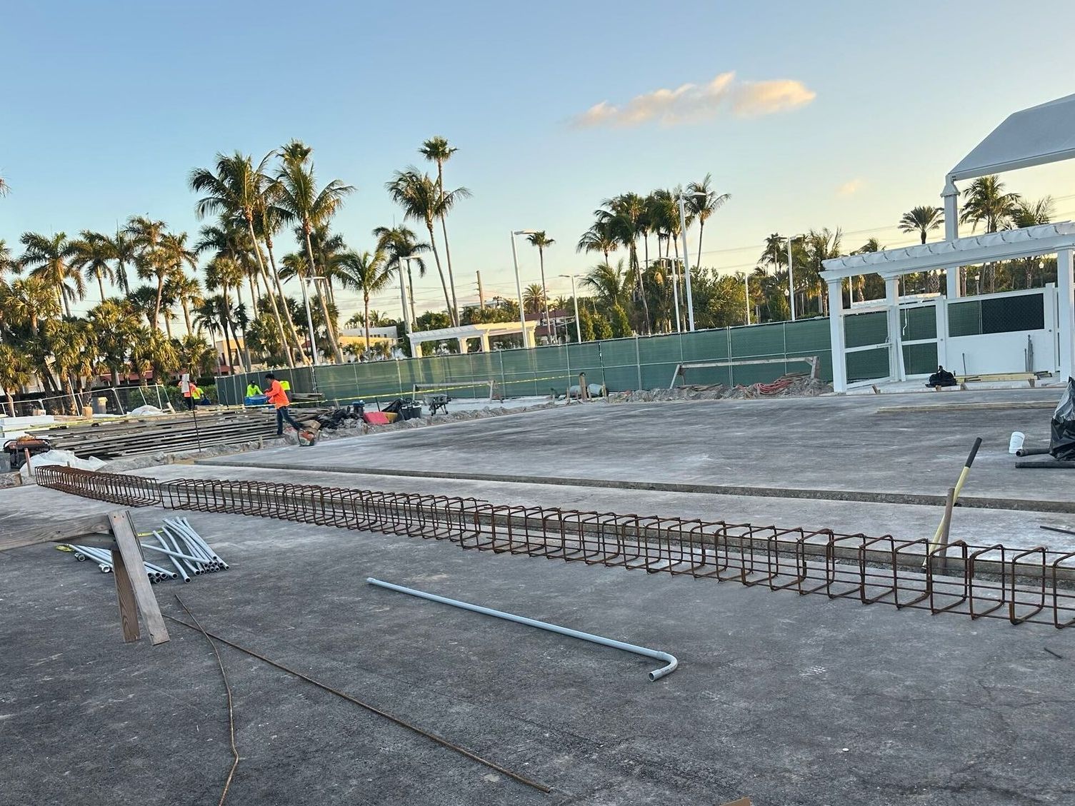 Beach construction site with rebar forms, concrete slabs, palm trees, and a white building at sunset
