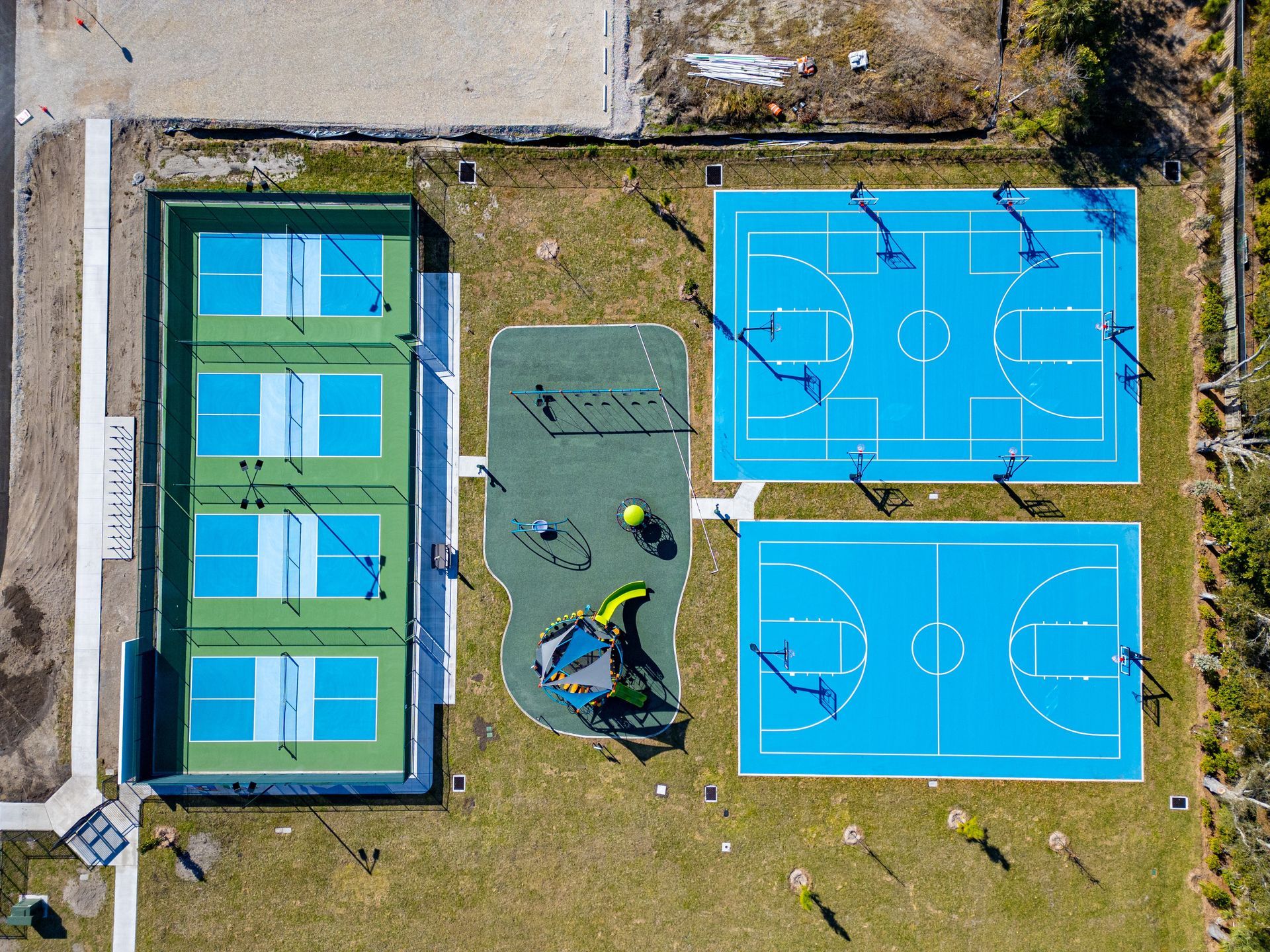 Aerial view of a sports complex with a tennis court, two blue basketball courts, and a small playground.