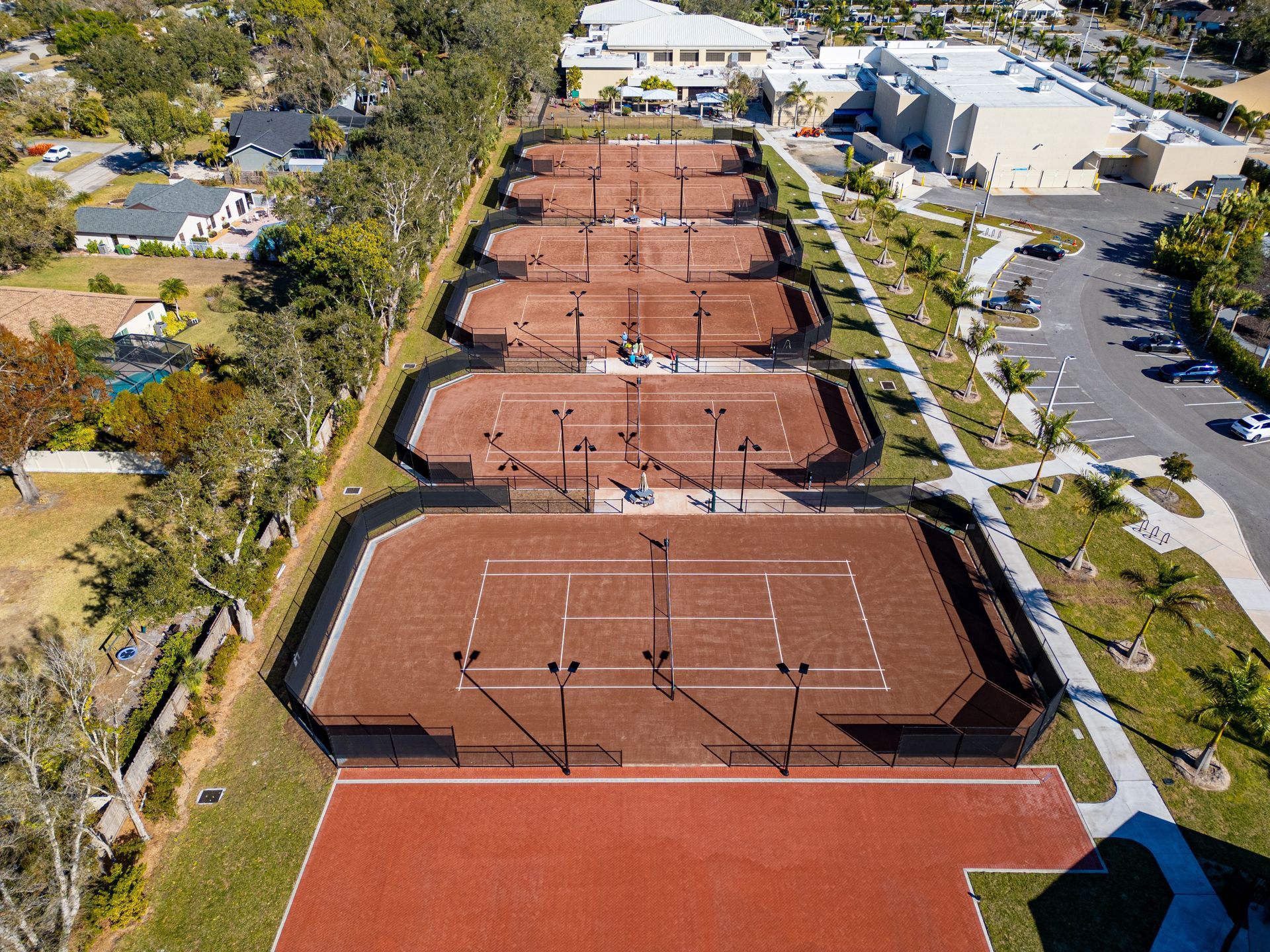 Aerial view of brown tennis courts beside a parking lot and trees.