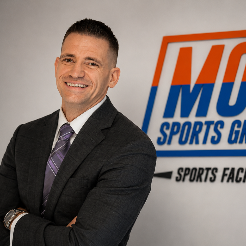 Keith Freie - Man in dark suit standing beside MOR Sports Group Sports Facilities sign, arms crossed and smiling