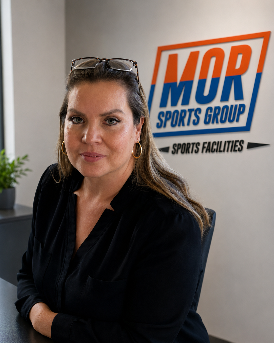 Woman standing in an office beside a MOR Sports Group sign, wearing a black blazer and glasses on her head